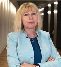 A woman with blonde hair wearing a light blue blazer and a blue top, standing with arms crossed in an indoor setting with bright lights.