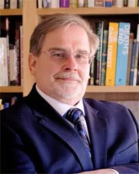 A middle-aged man with glasses, gray hair, and a beard, wearing a navy blazer and tie, standing in front of a bookshelf filled with books.