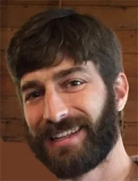 A smiling man with dark hair and a beard, indoors with a wooden background.