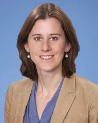 Professional woman with shoulder-length brown hair, wearing a beige blazer and blue top, smiling against a blue background.