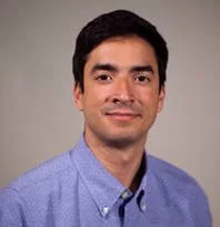A portrait of a young man with dark hair, wearing a light blue collared shirt, smiling slightly against a plain gray background.