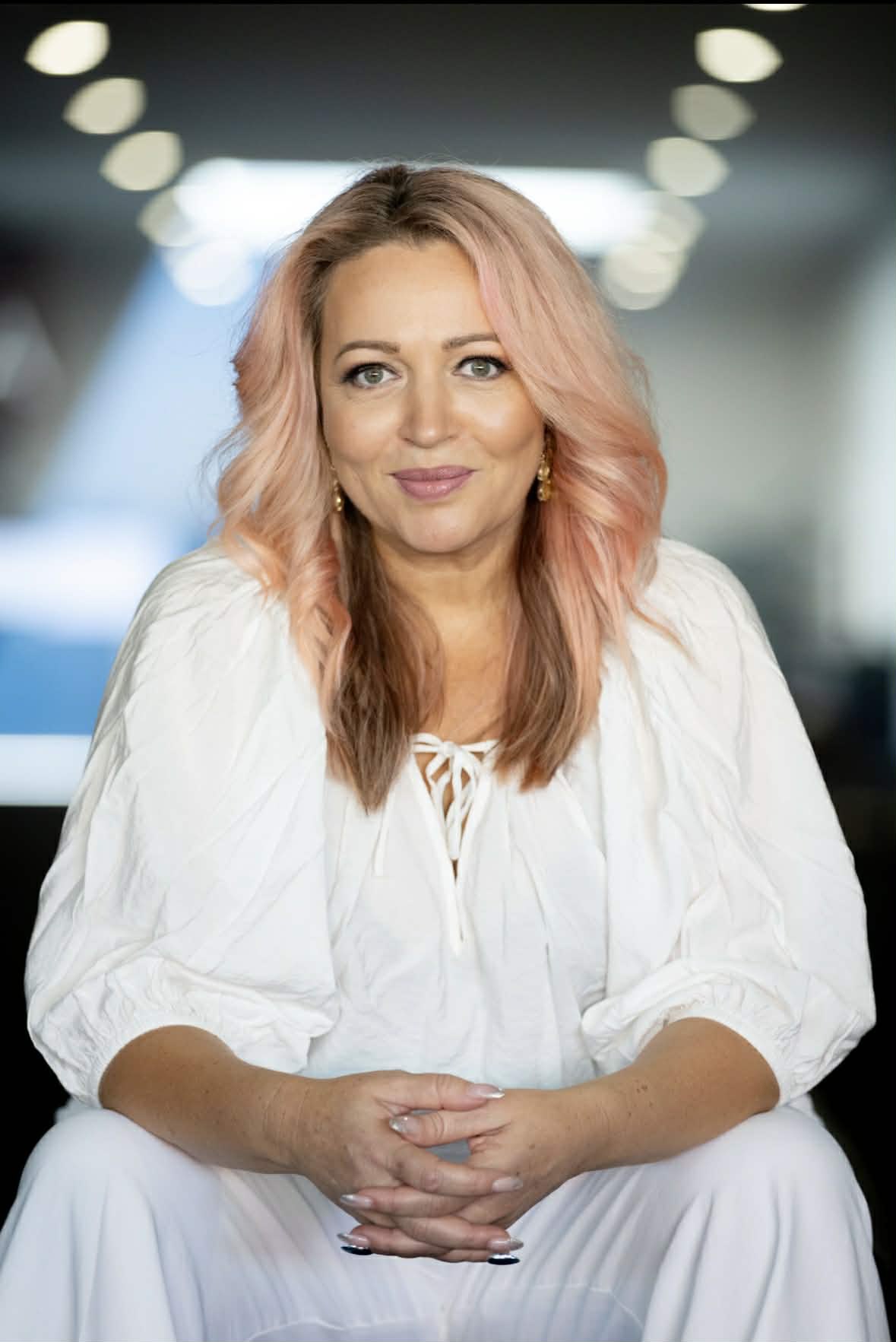 A woman with pinkish blonde hair and light skin, wearing a white top, sitting indoors with a blurred background of blue and gray tones. She is smiling slightly and looking at the camera.