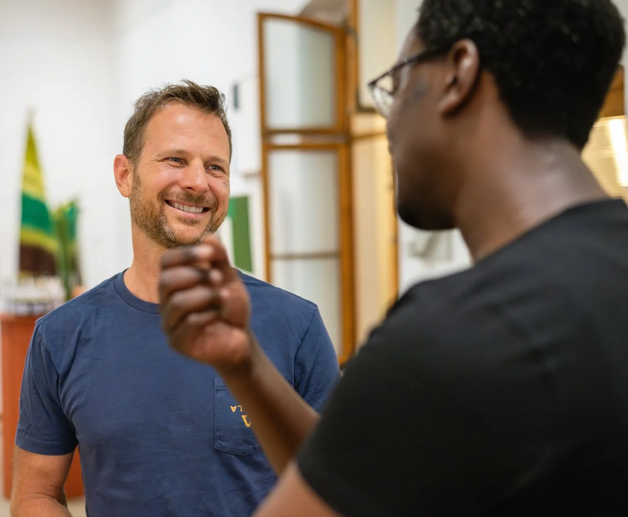 Two men smiling and having a conversation indoors, one facing the camera with a beard and blue shirt, the other facing away with glasses and black shirt.
