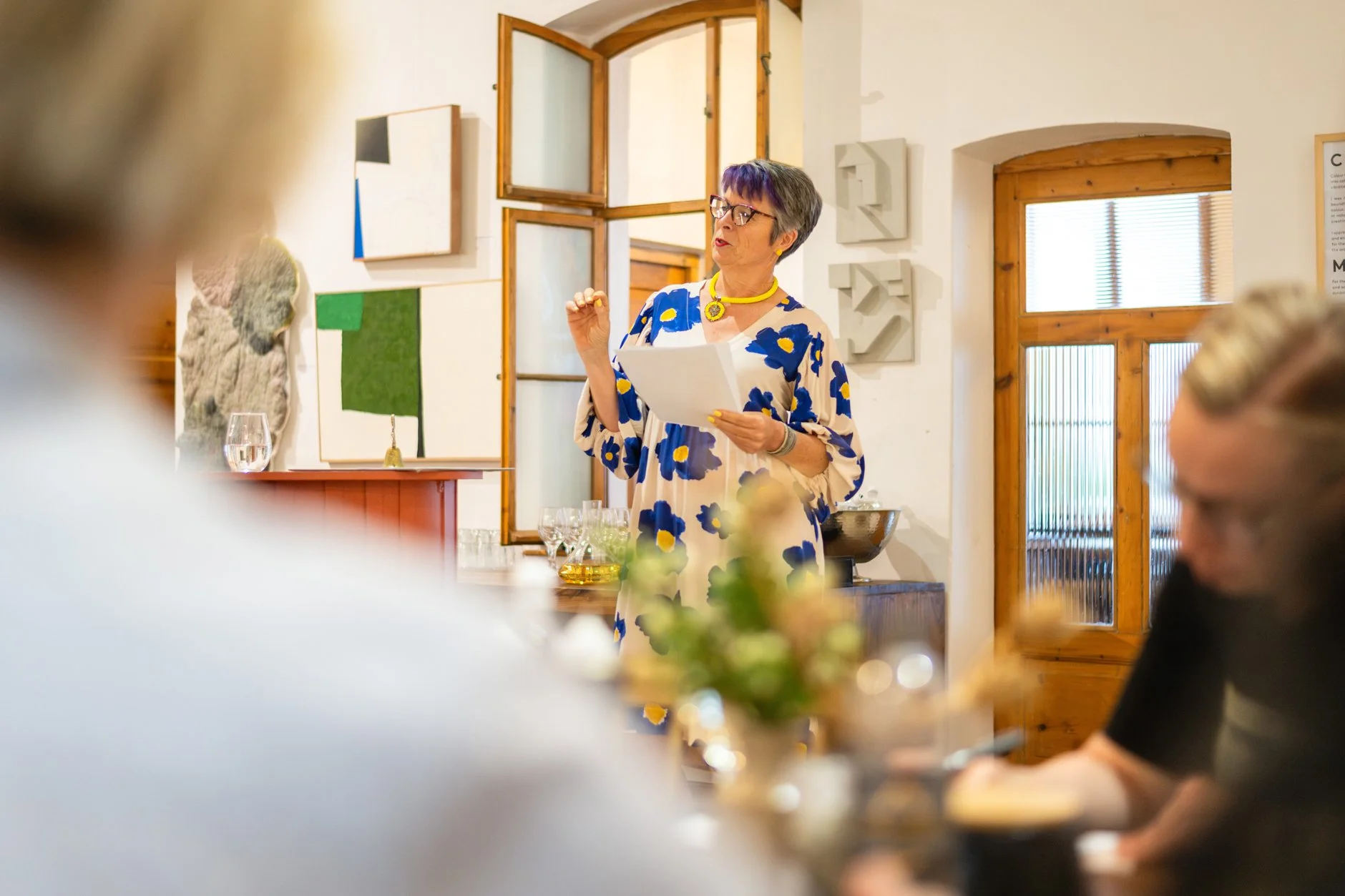 A woman in a blue and white floral dress speaking at a gathering during the daytime. She holds a paper and gestures with her hand. The room has artwork on the walls, windows with wooden framing, and a table with glasses and a flower arrangement in the foreground.