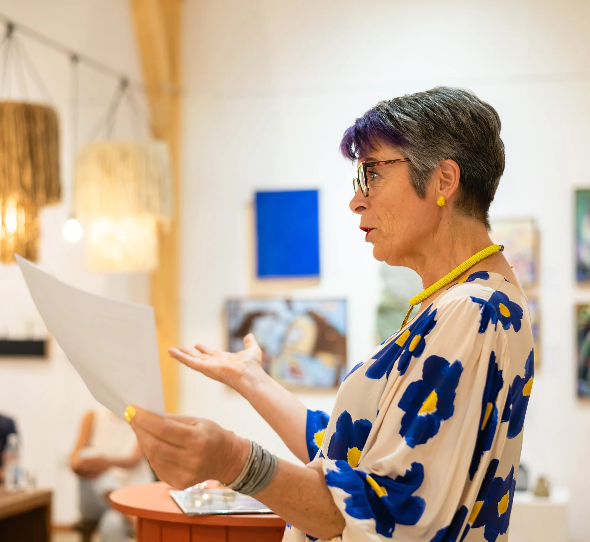 A woman with short gray hair and glasses, wearing a beige dress with blue and yellow flowers, reading a piece of paper in an art gallery.