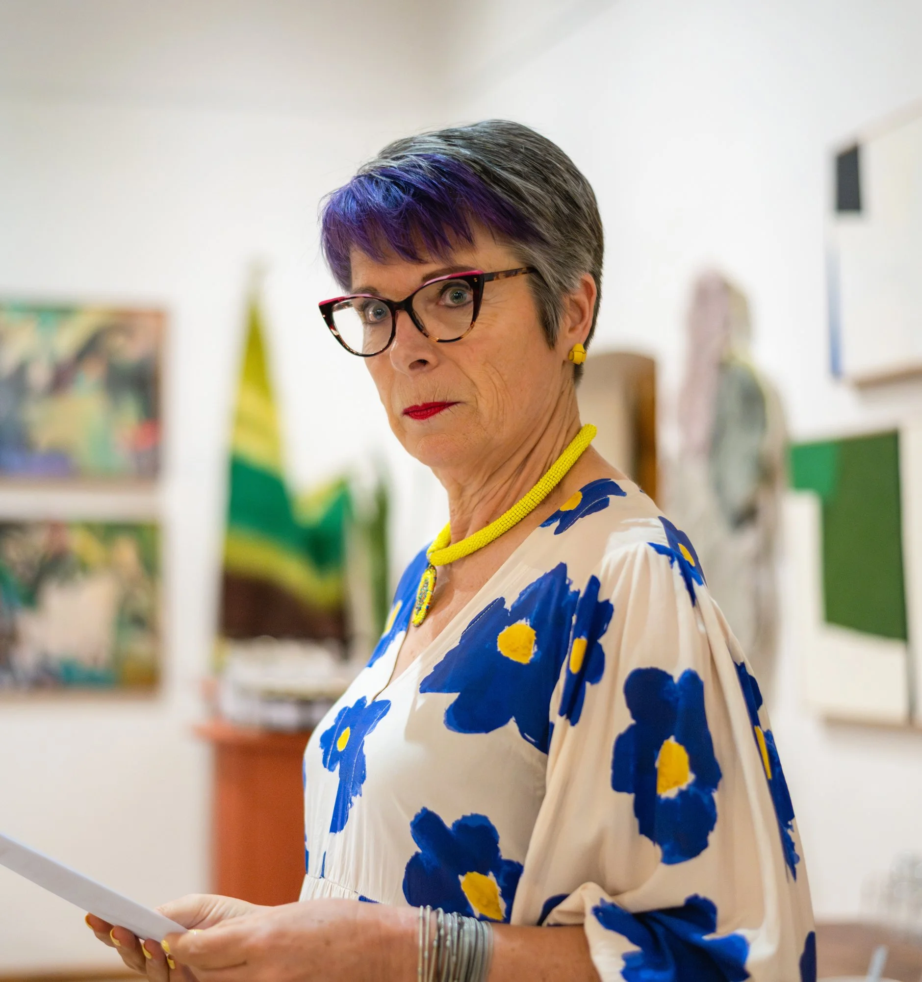 A woman with short gray hair and glasses, wearing a floral blouse with blue and yellow flowers, a yellow necklace, and yellow earrings, standing indoors with art and a green flag in the background, holding a piece of paper and looking at the camera.