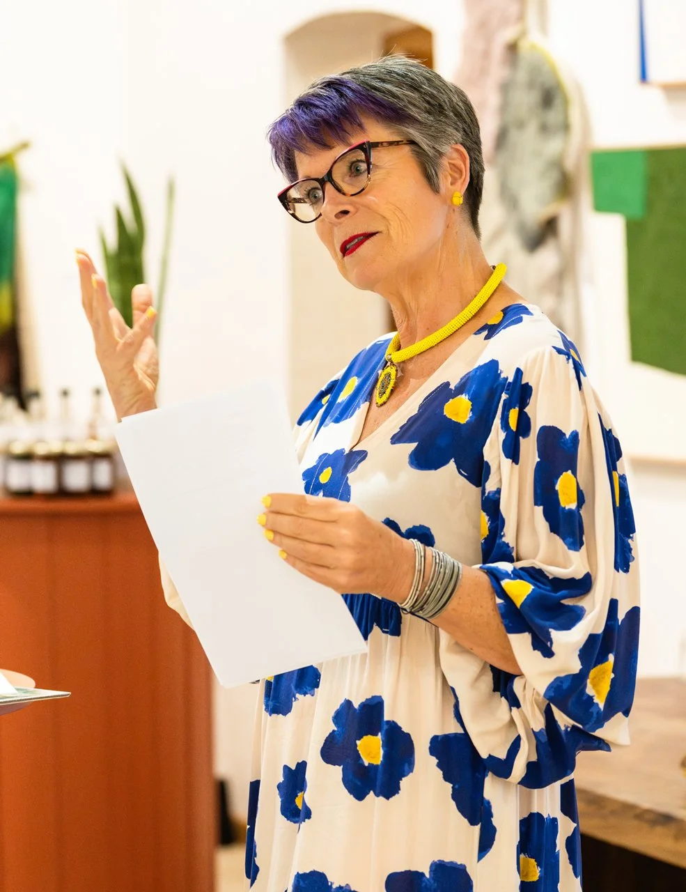 An older woman with short gray hair, wearing glasses, large yellow jewelry, and a white dress with blue and yellow floral pattern, holding a piece of paper and gesturing with her hand in an indoor setting.