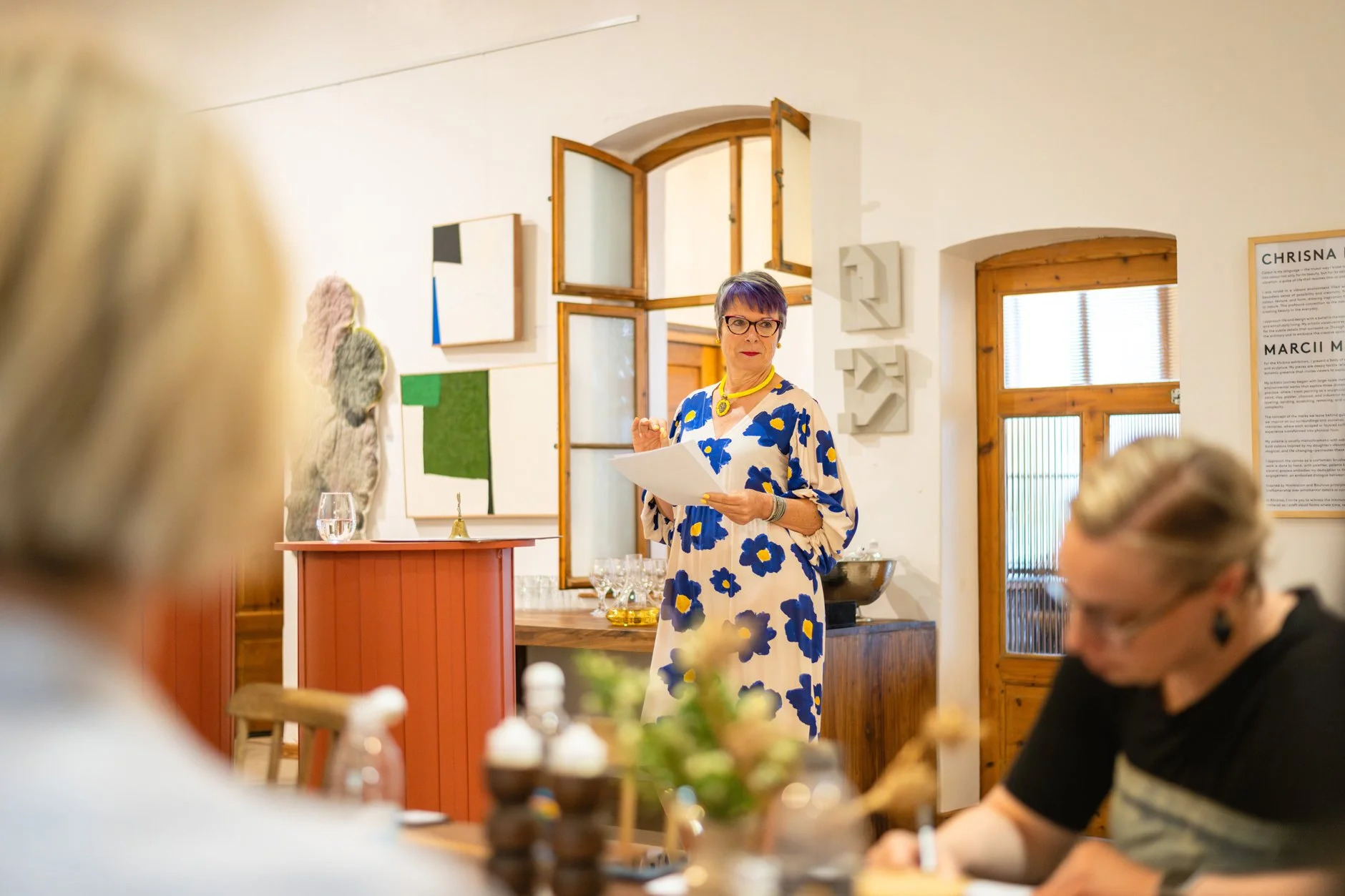 A woman wearing a floral dress and glasses stands in a room, holding papers, during a presentation or speech. There are artworks on the wall behind her, and two women are seated at a table in the foreground.