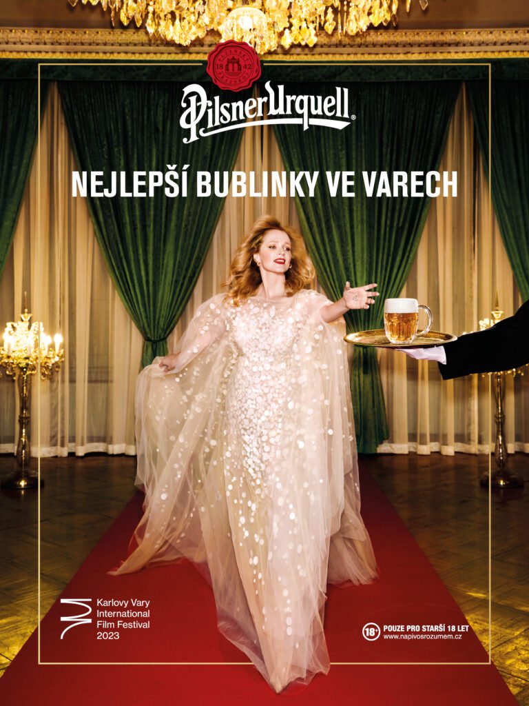 A woman in an elegant, sparkling gown walks down a red carpet in a decorated room with green curtains and gold accents, holding her arm out as if reaching for a drink served by a person in a black suit. There is a chandelier hanging from the ceiling. The scene appears to be an event at the Karlovy Vary International Film Festival in 2023.