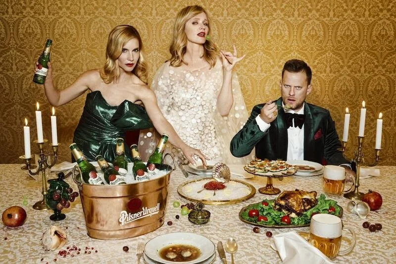 Three people dressed formally at a dinner table with candles, food, drinks, and decorations, in front of a patterned gold wall.