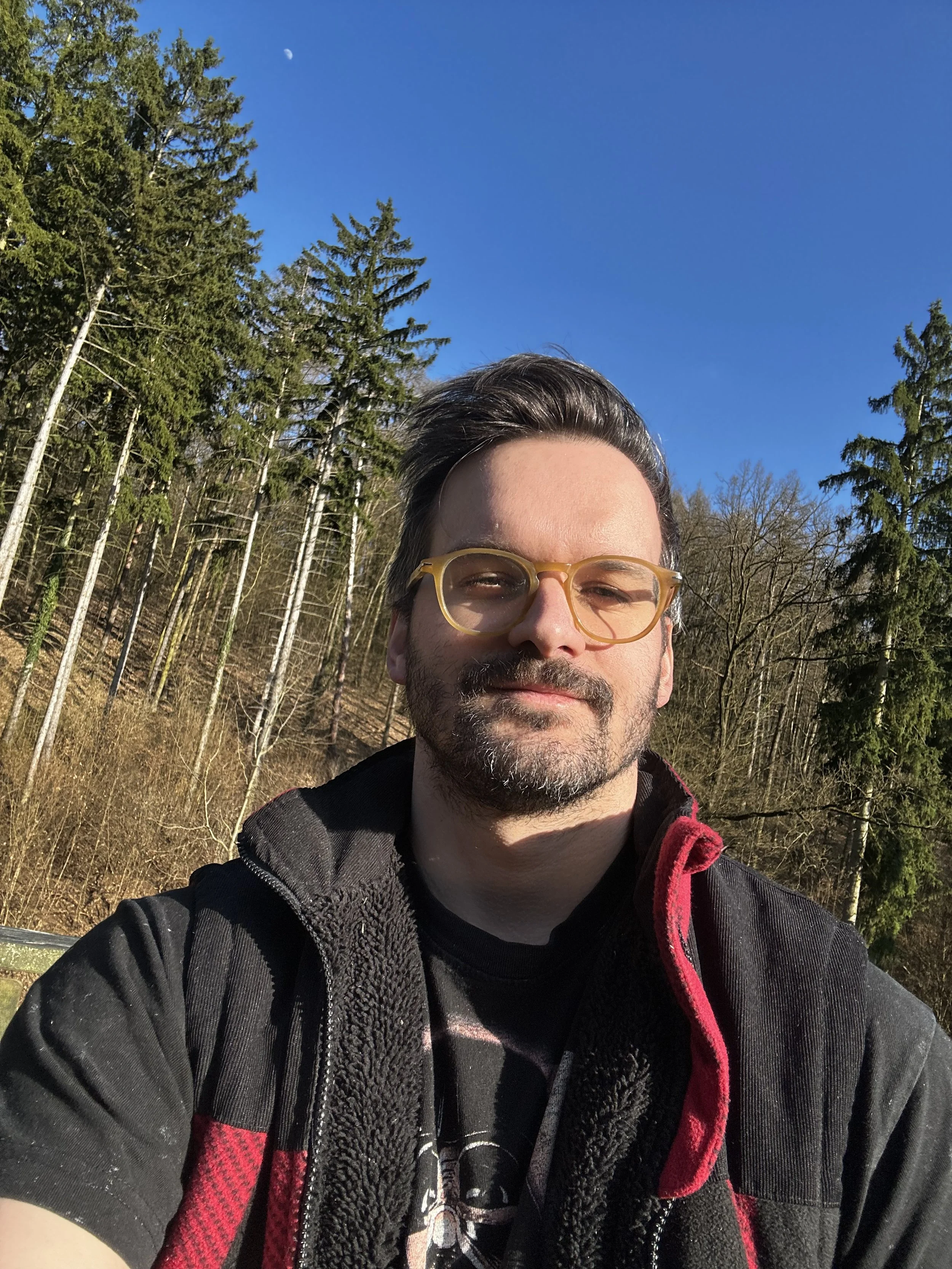 A man with glasses taking a selfie outdoors. He is wearing a black and red jacket and has short dark hair with some gray. The background includes tall evergreen trees, leafless trees, and a clear blue sky with a visible moon.