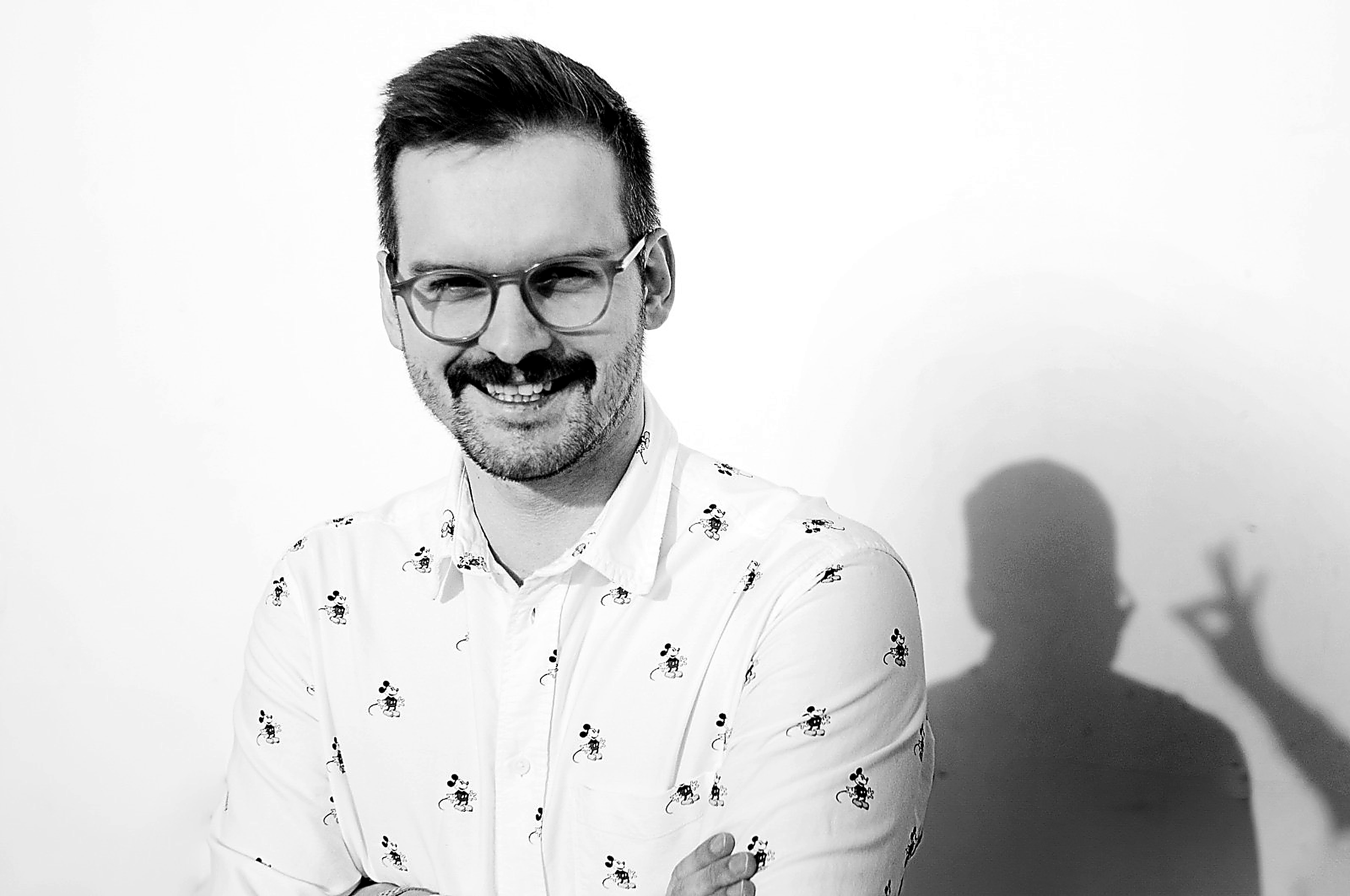 A black-and-white photo of a young man with glasses, a beard, and a mustache, smiling and looking to the side. He is wearing a white shirt with small Mickey Mouse print and crossing his arms, standing against a plain white background.