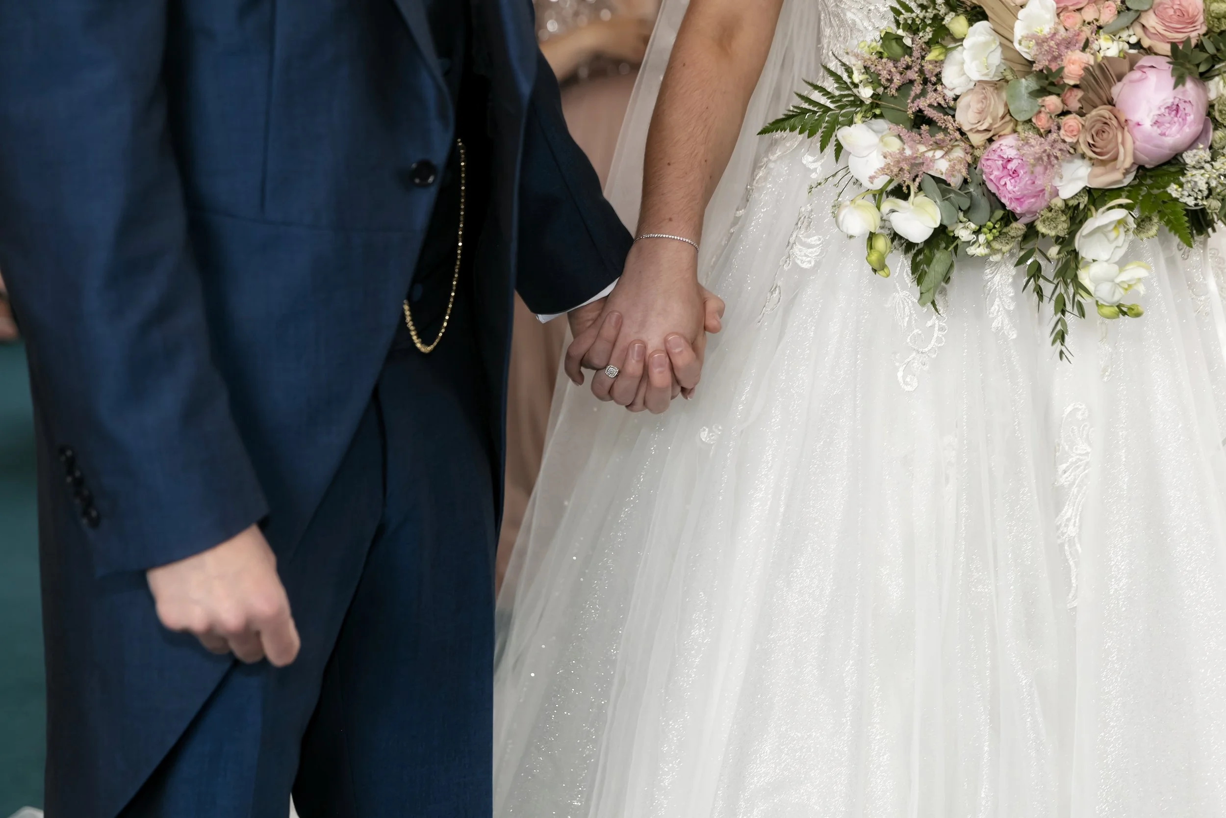 Close-up of a bride and groom holding hands during their wedding ceremony, with the bride wearing a white dress and floral bouquet, and the groom dressed in a dark suit.