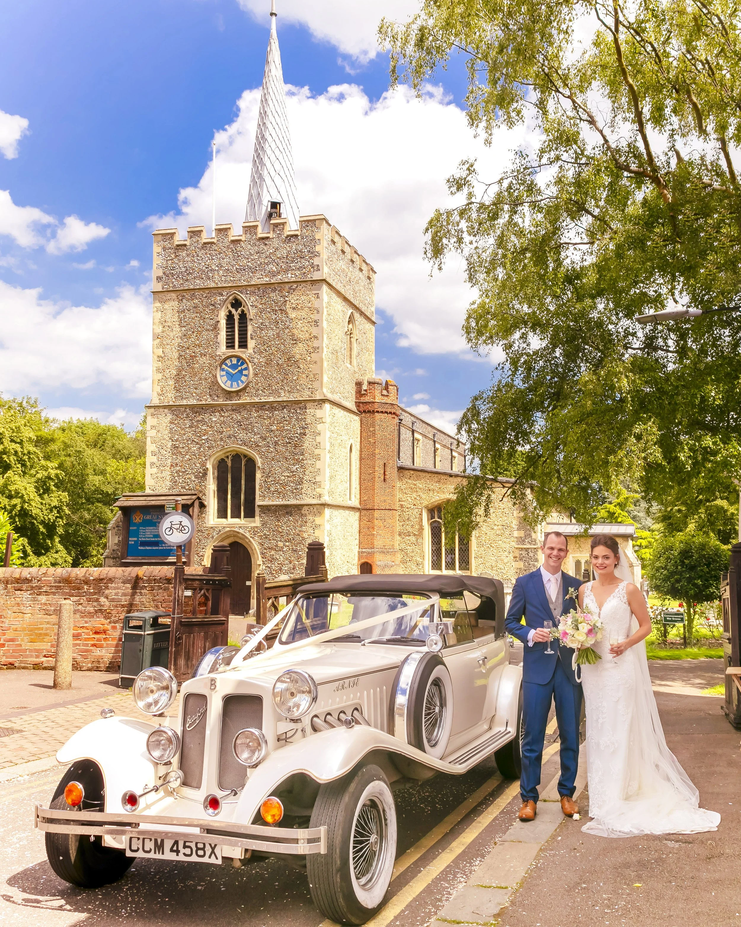 A bride and groom standing next to a vintage car outside a church, with trees and a blue sky in the background.