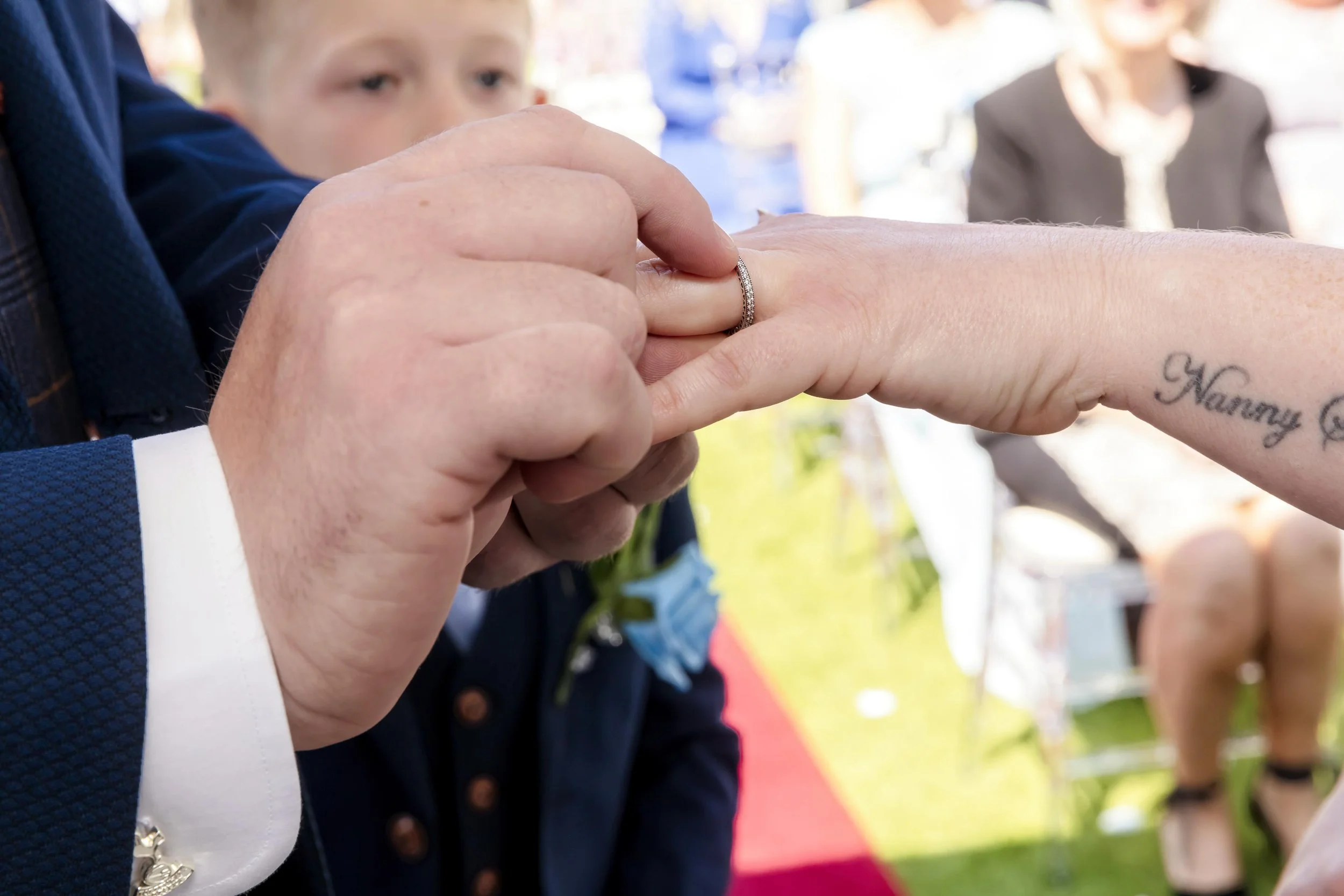 A man is placing a ring on a woman's finger during a wedding ceremony outdoors, with a young boy and a woman in the background.