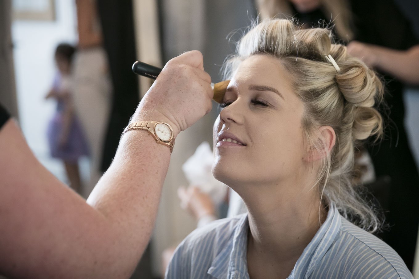 A woman with blonde hair in curlers having her makeup done by a makeup artist, with a blurred person in the background.