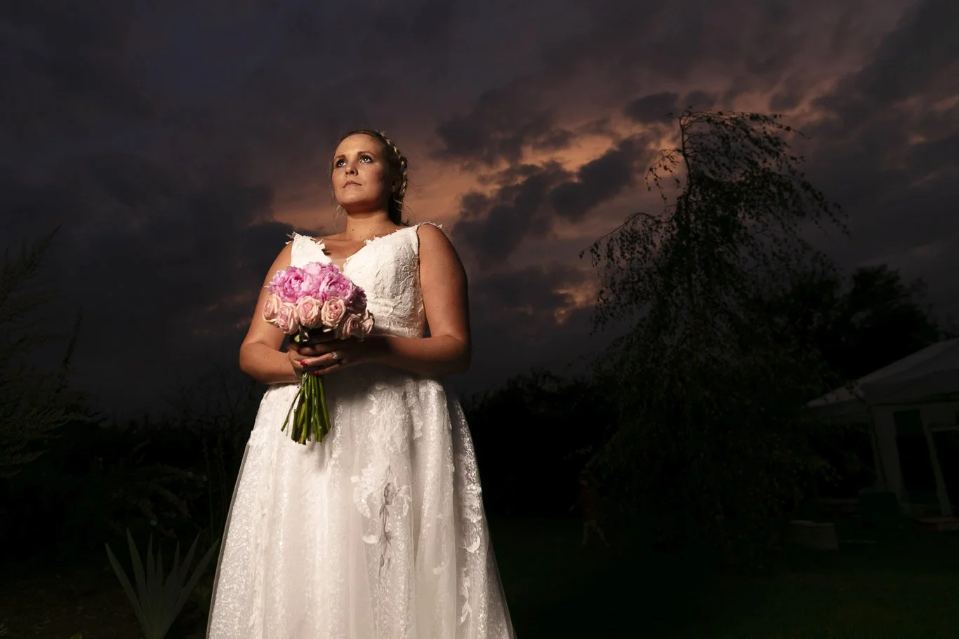 A bride in a white lace wedding dress holding a bouquet of pink and cream roses, standing outdoors during dusk with a dark, cloudy sky in the background.