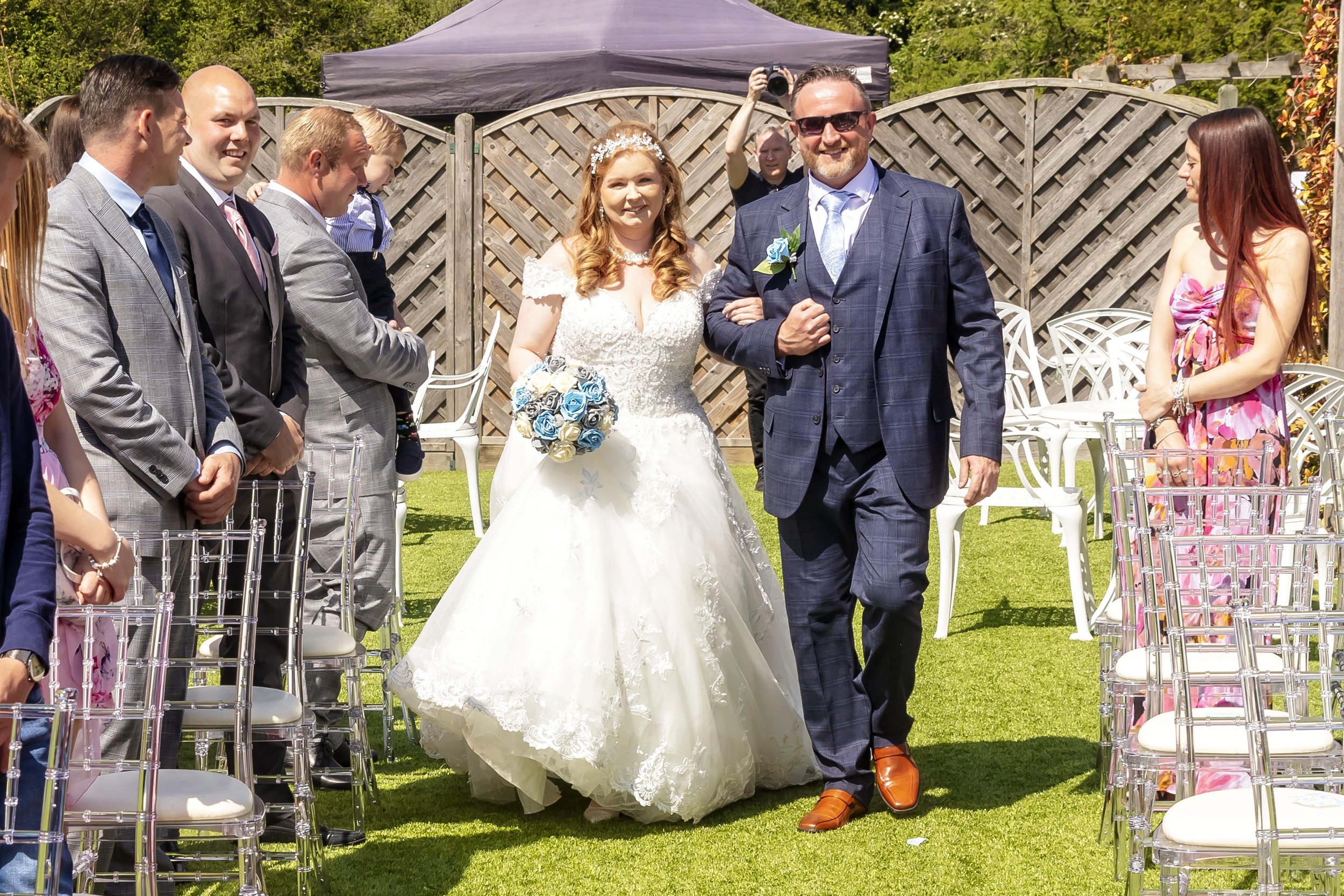 A bride in a white wedding dress holding a bouquet of blue roses walking down the aisle with a man, likely her father, in a blue suit. Guests are standing on either side of the aisle, smiling and watching. There is a black canopy tent in the backgrou