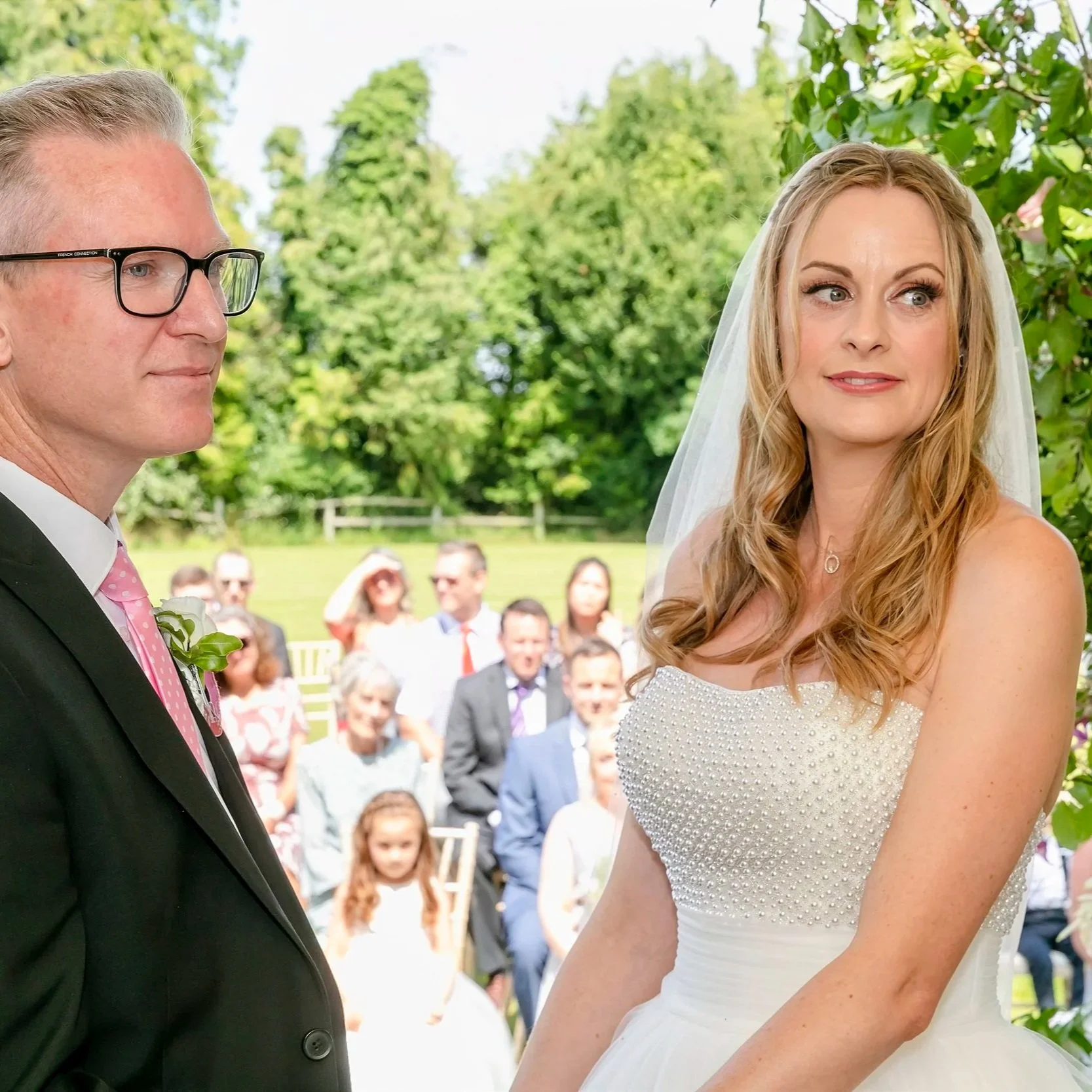 Bride and groom standing outdoors during wedding ceremony with guests in background.