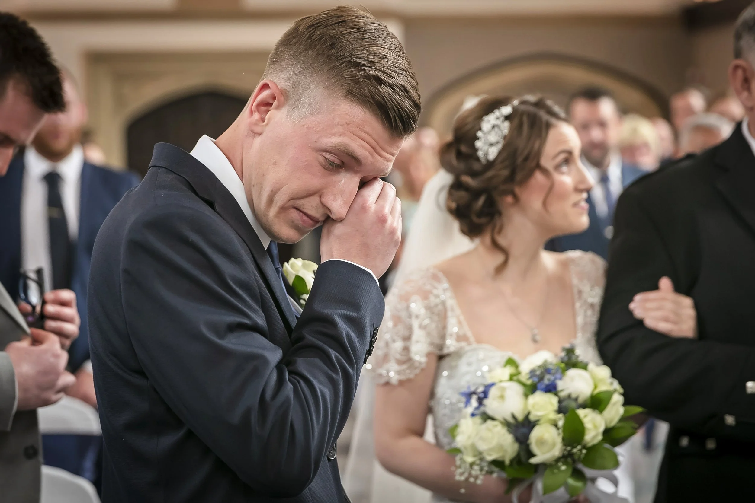 A groom in a dark suit with a white boutonniere is holding his face with one hand in emotional distress during a wedding ceremony. Beside him, a bride in a lace wedding dress with a hairpiece and veil holds a bouquet of white roses and blue flowers, 