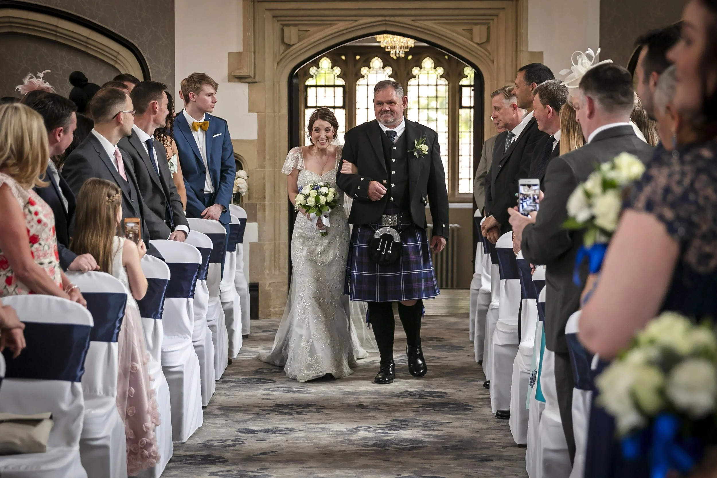 Bride walking down the aisle with her father at a wedding ceremony in a church, surrounded by seated guests taking photos and videos.