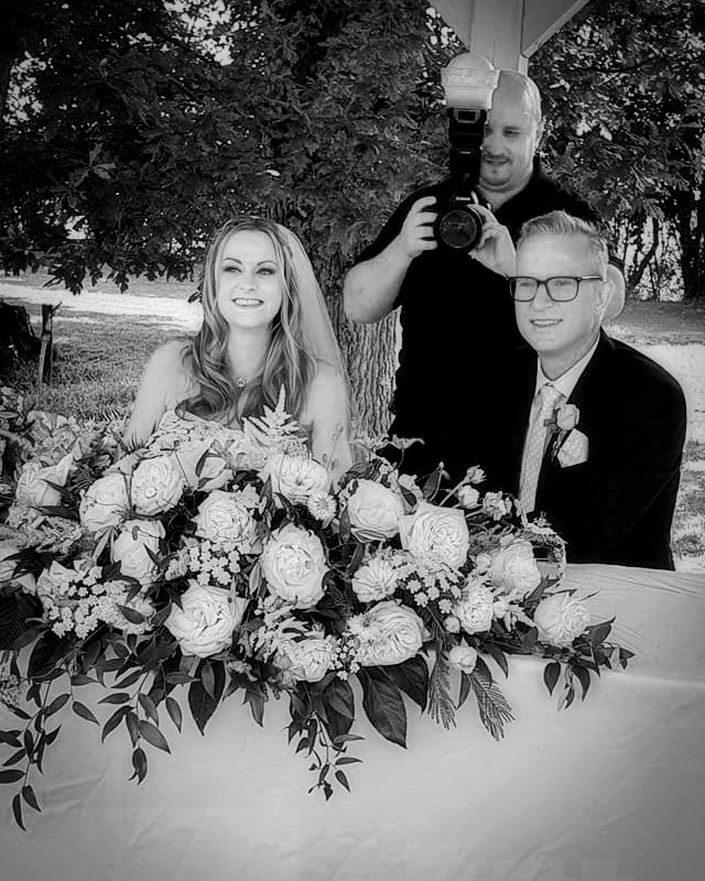 A wedding or special event scene with a woman and man seated in front of a large floral arrangement featuring roses and greenery, with a photographer taking their picture outdoors under a tree.