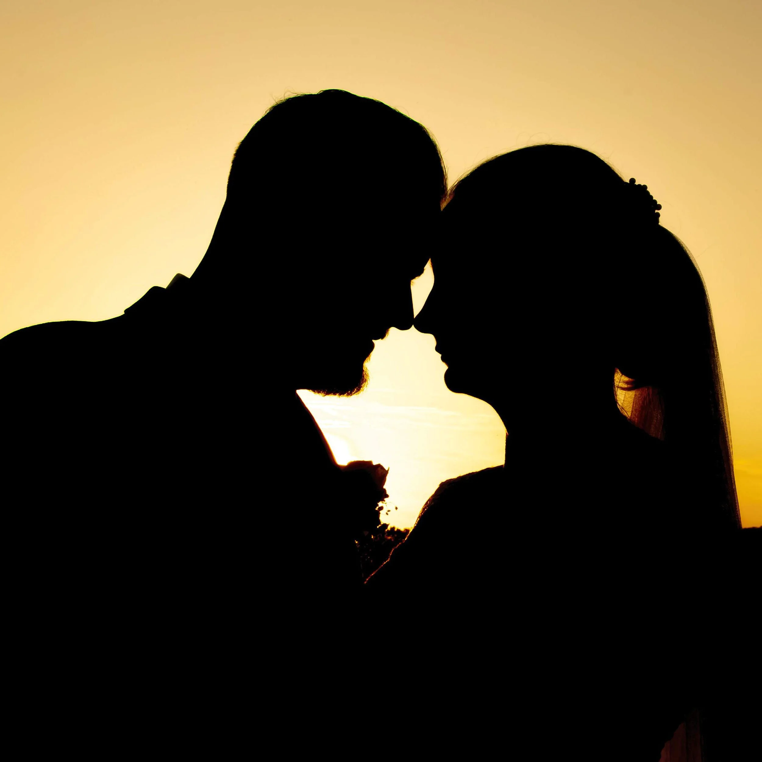 Silhouette of a couple touching foreheads at sunset, with warm yellow and orange sky in background.