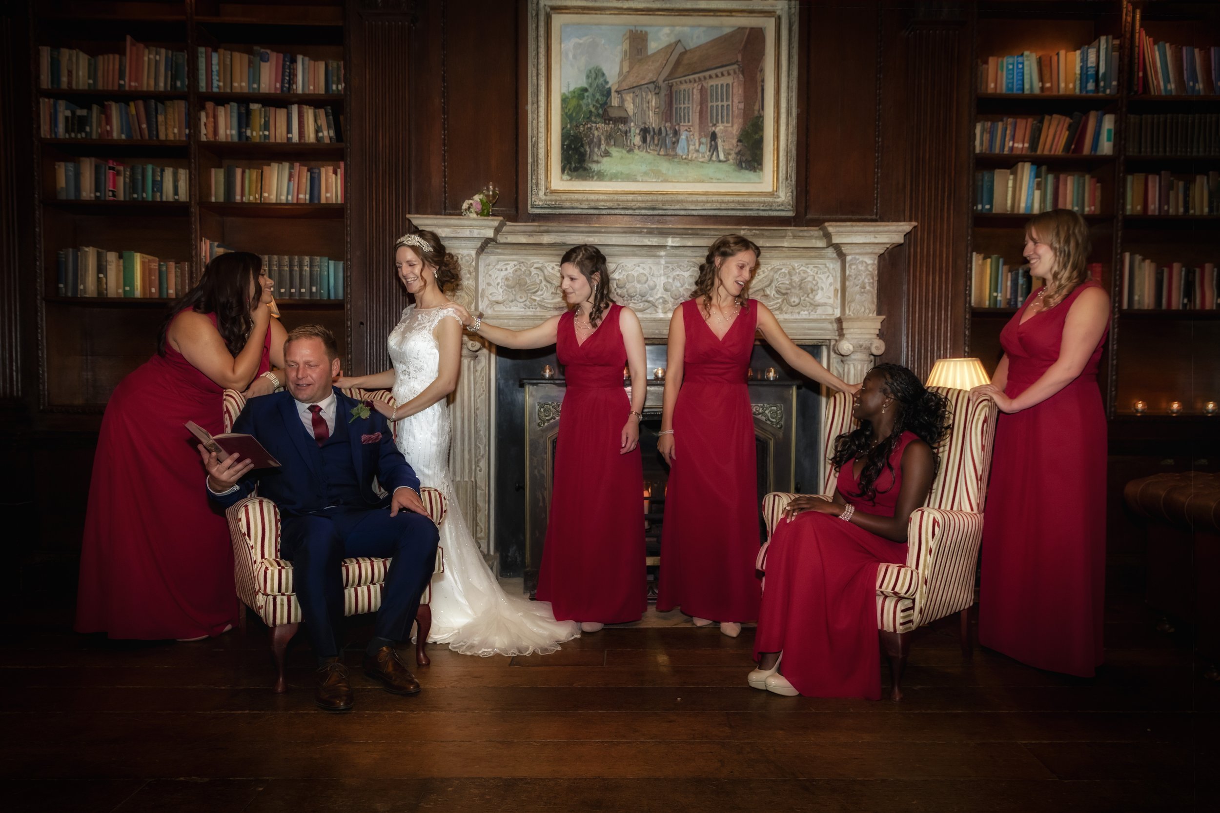 A bride and groom with six bridesmaids in red dresses and a woman in a burgundy gown, posing in a wood-paneled room with bookshelves, fireplace, and a painting, during a wedding celebration.