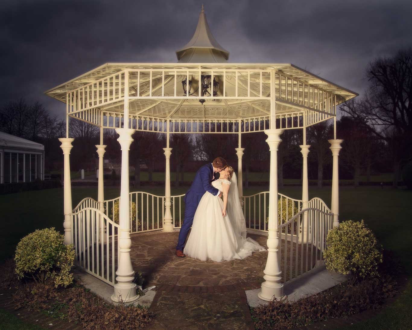 A bride and groom in wedding attire sharing a romantic moment under a white gazebo outdoors during dusk, with dark sky and trees in the background.