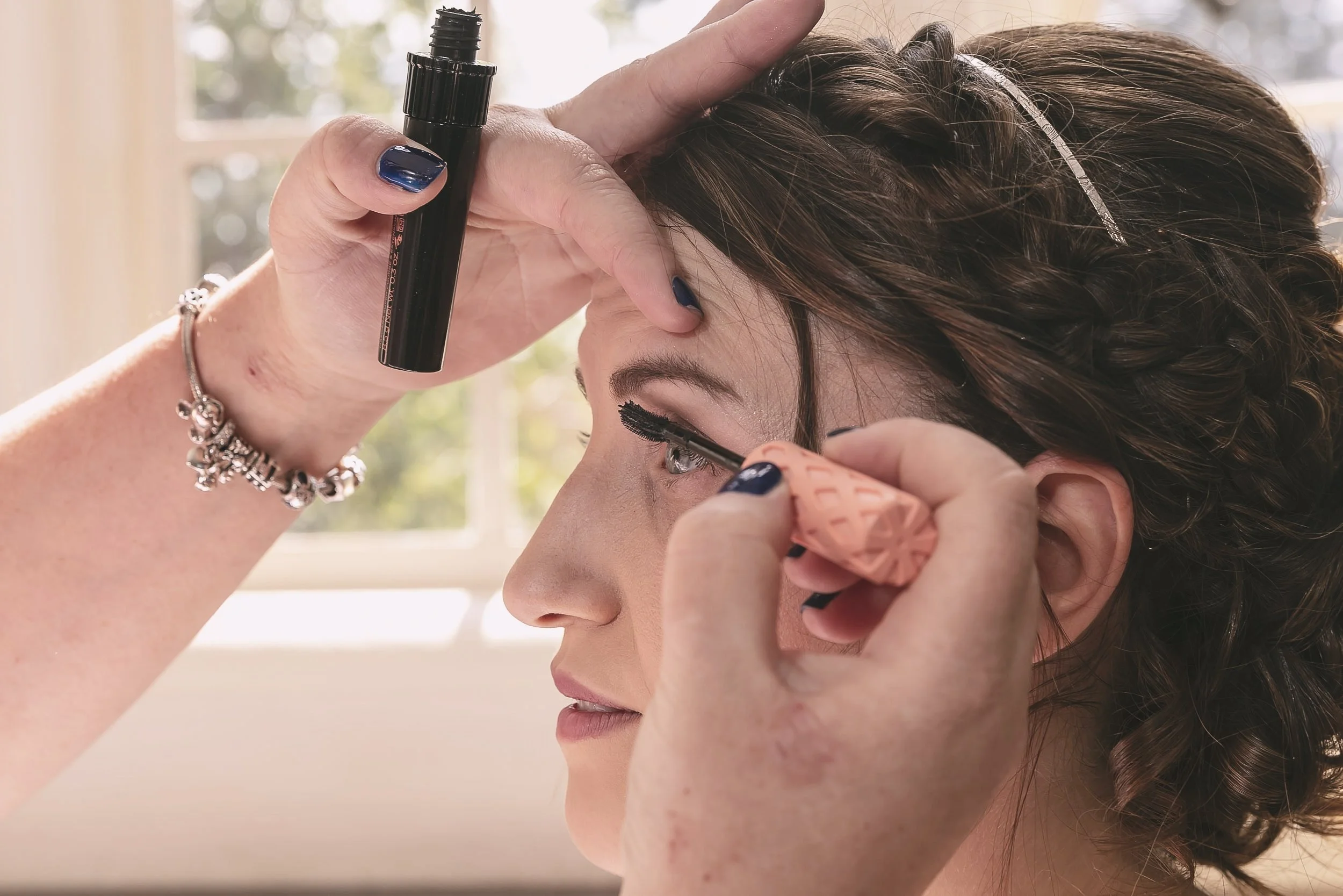A woman is getting her eyelashes done. A beautician is applying black mascara or eyelash extension adhesive to her upper eyelashes using a small brush, while holding her eyelid. The woman has curly brown hair and is wearing a sparkly headband. She is sitting near a window with a blurred outdoor background.