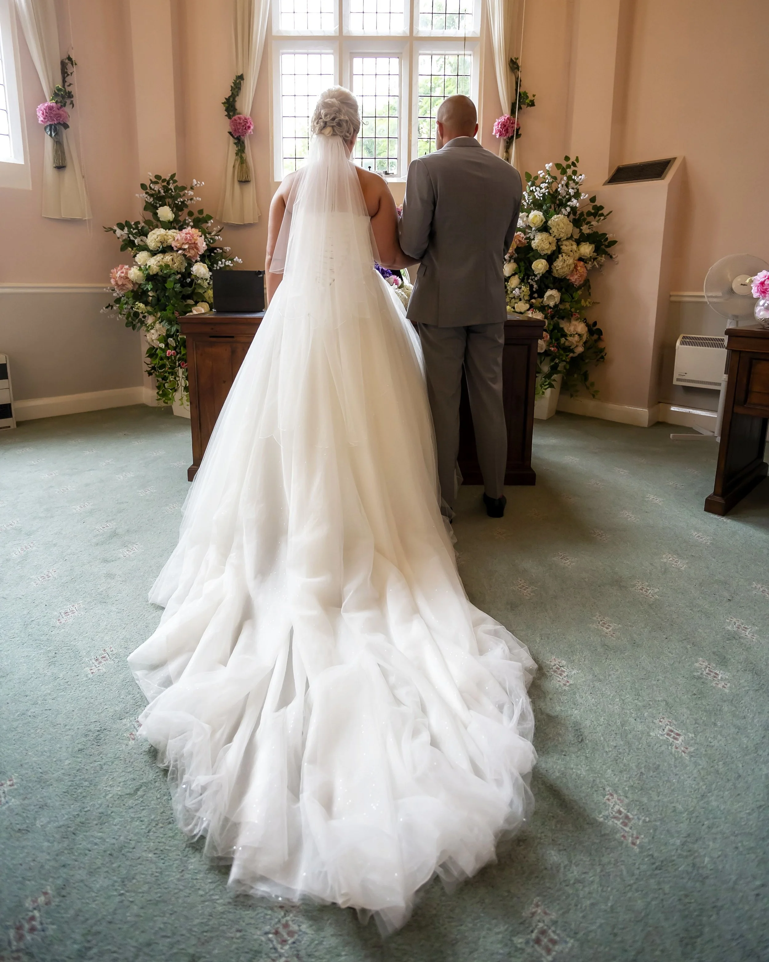 A bride and groom standing side by side during their wedding ceremony in a softly lit room with floral decorations on the wall and a window behind them.