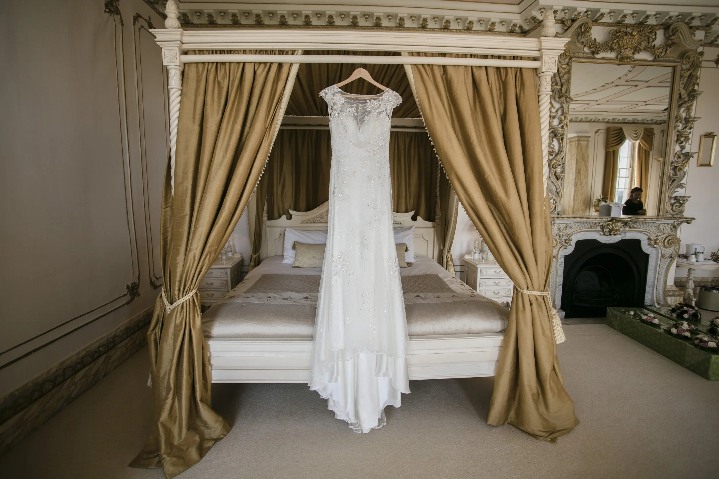 A wedding dress hanging from a wooden hanger on a bed's four-poster canopy, with gold curtains, in an ornate bedroom with a large mirror.