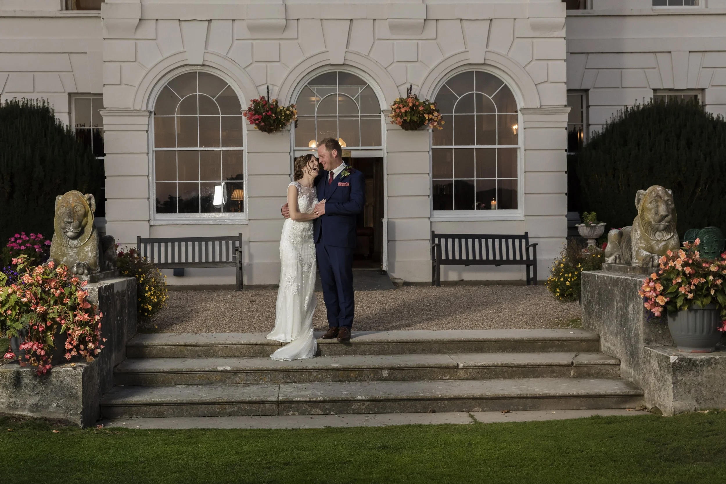 A newlywed couple in wedding attire stands on steps outside a white building with large arched windows, surrounded by flowers and lion statues.
