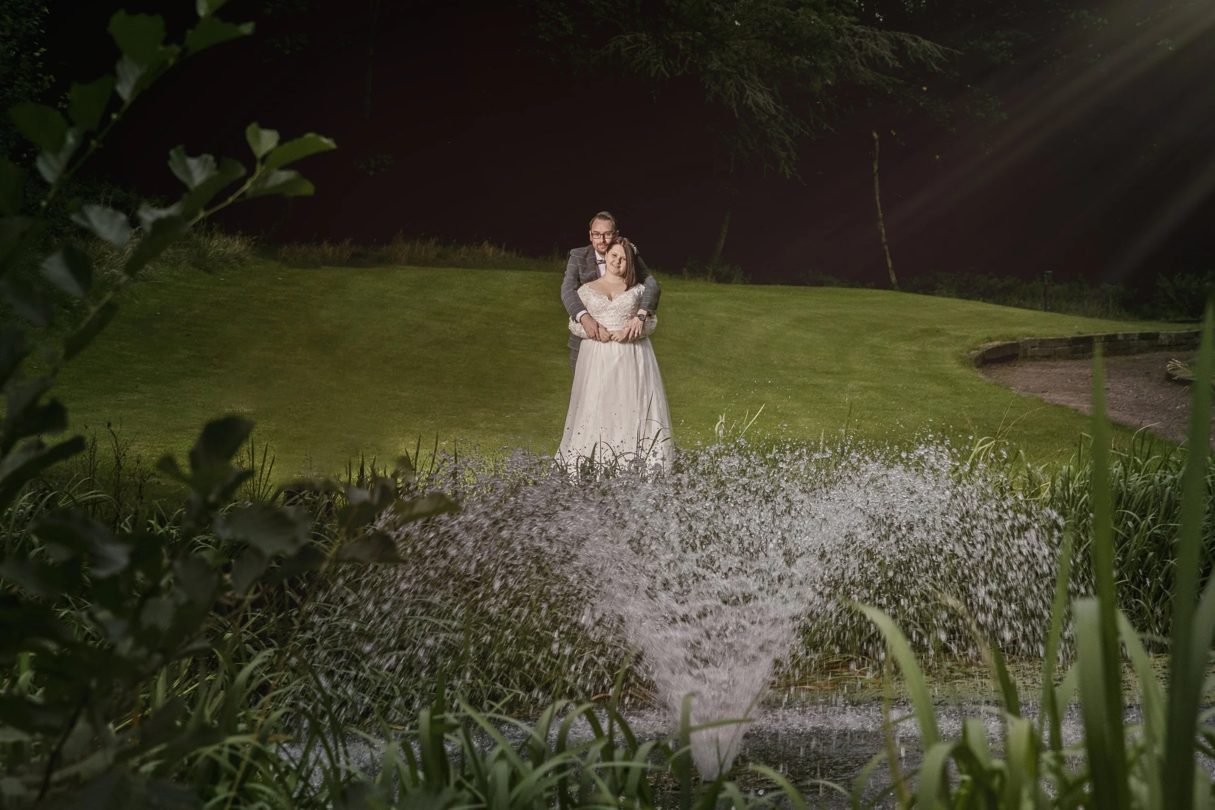 A bride and groom at night, standing on a grassy area near a pond with water spray in the foreground, surrounded by greenery and illuminated by a light beam.