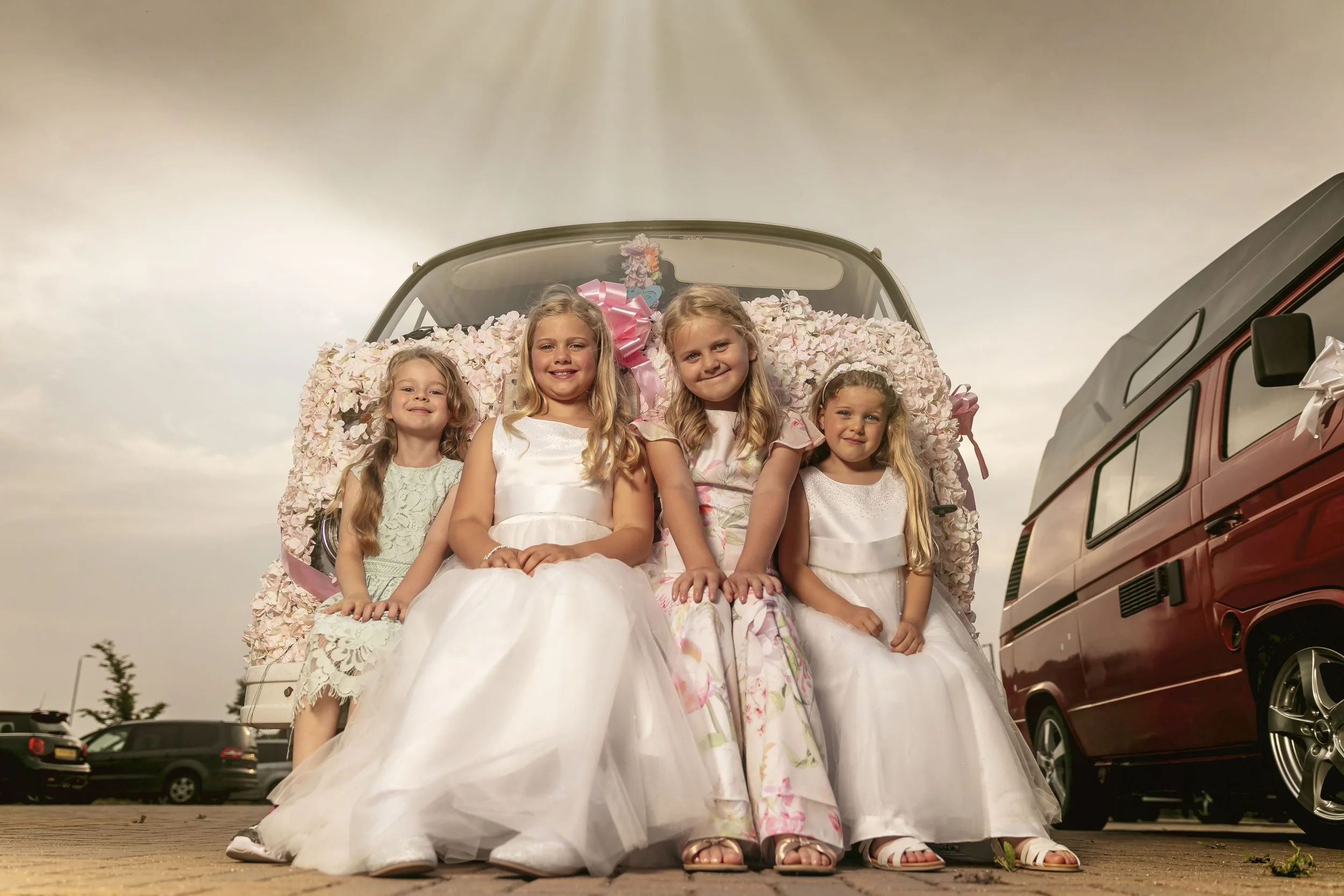Four young girls in fancy dresses sitting on the back of a decorated flower-covered vehicle, with two additional vehicles parked in the background under a cloudy sky.