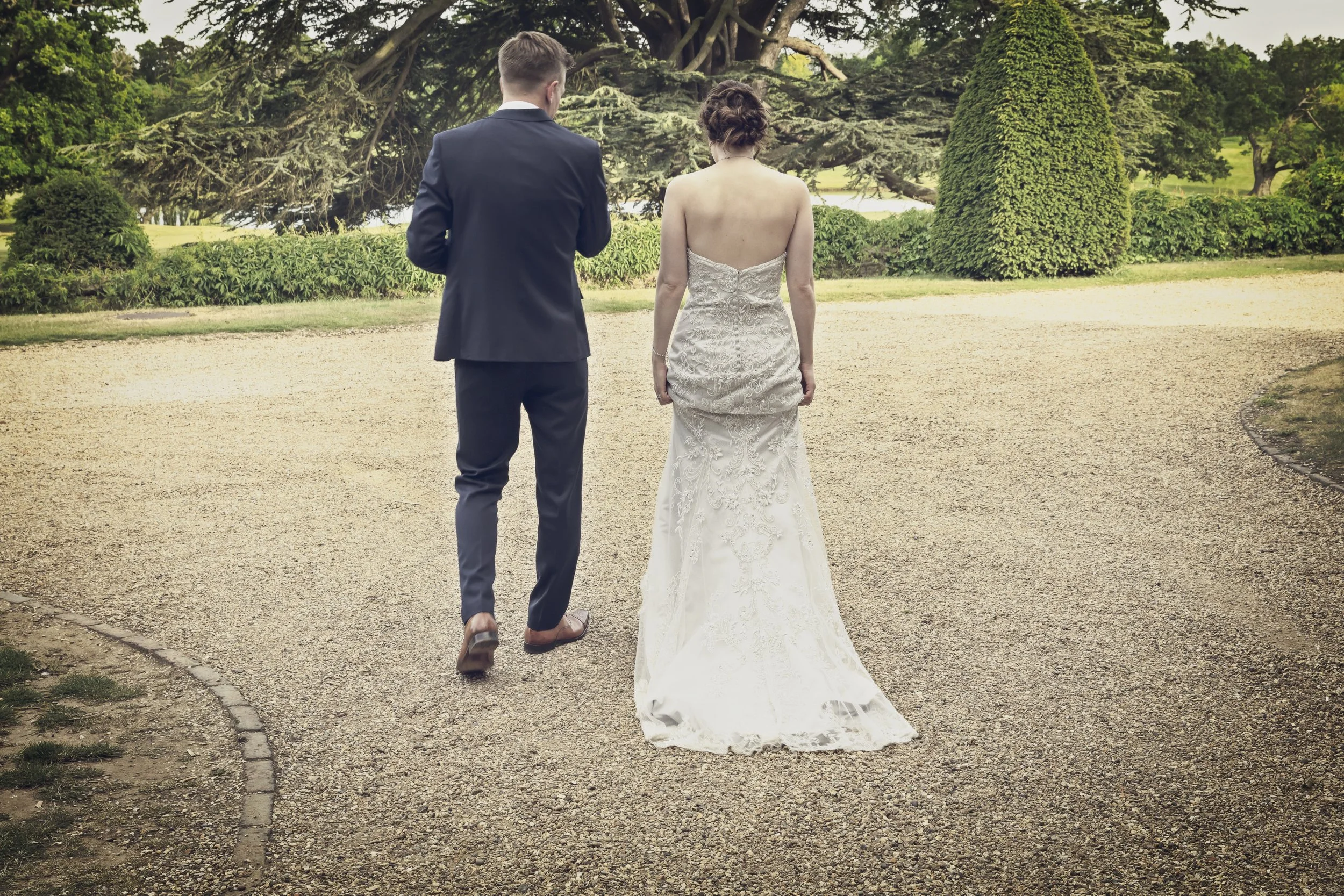 A bride and groom walking away on a gravel path in a park with green trees and shrubs.