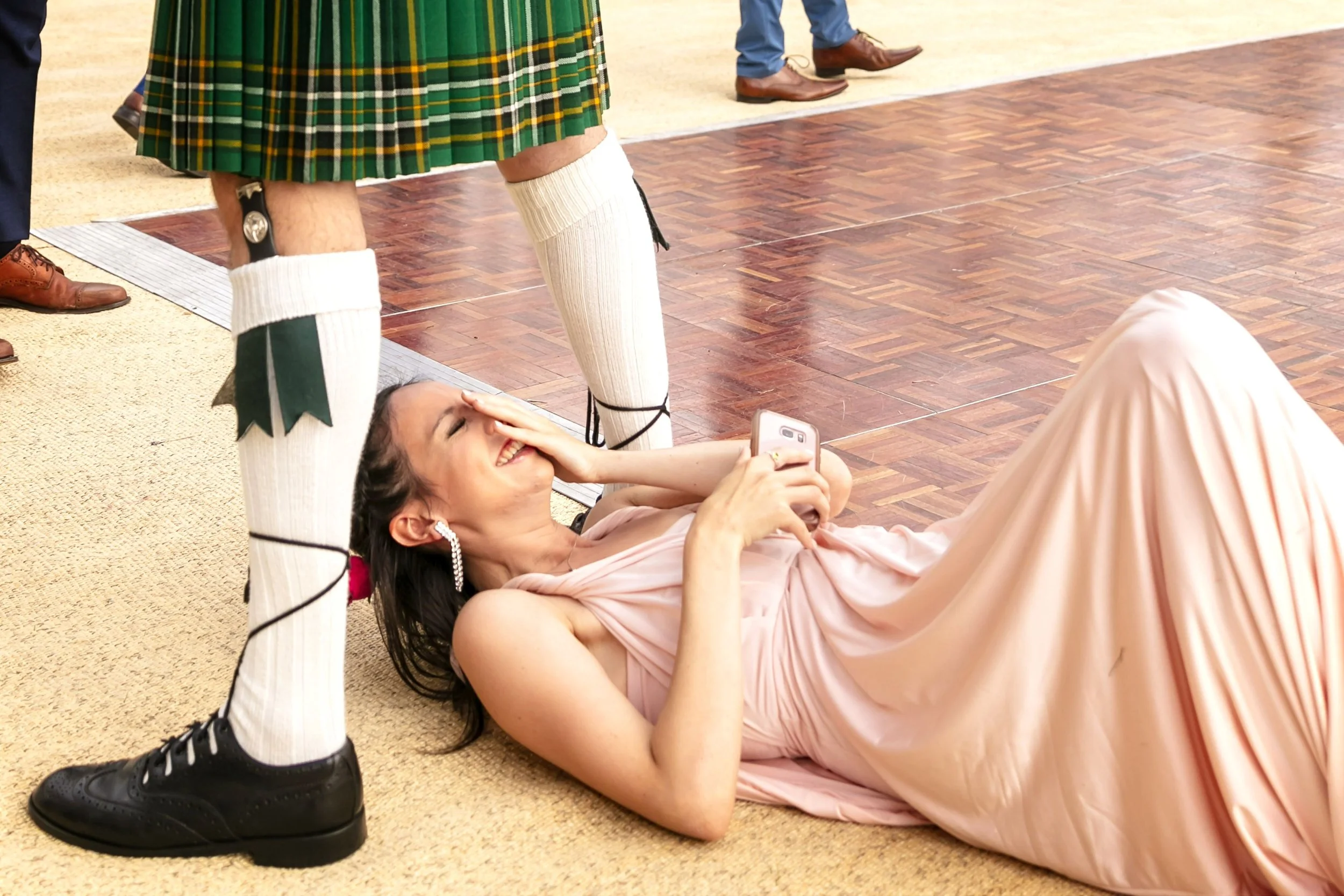 A woman lying on the floor, smiling and holding a phone, with a man standing over her wearing traditional Scottish attire, including kilt, white knee-high socks, and black shoes. Other people are visible in the background.
