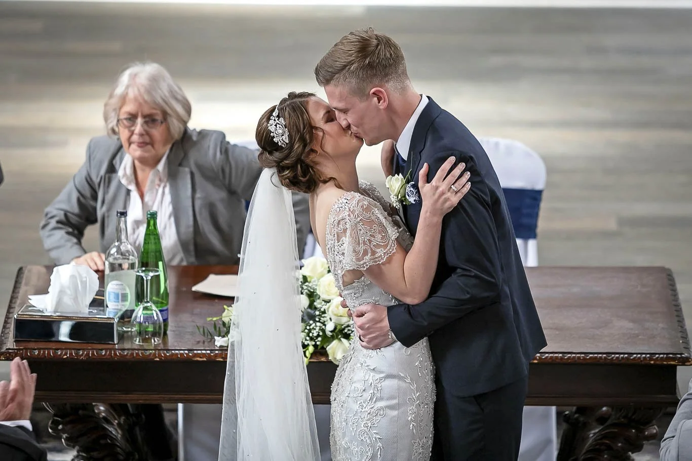 A newlywed couple sharing a kiss during their wedding ceremony, with an officiant sitting at a table nearby.