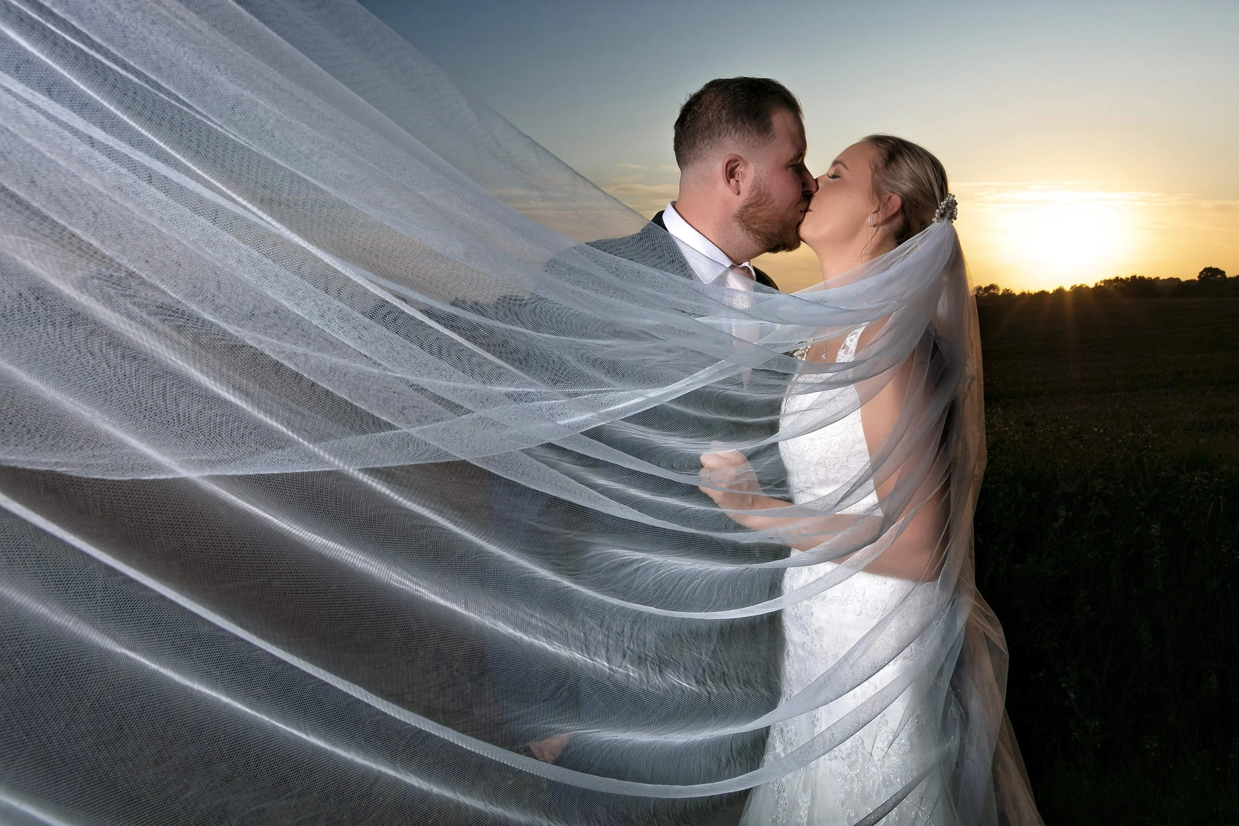 A bride and groom kiss outdoors at sunset, with the bride's veil flowing in the wind.