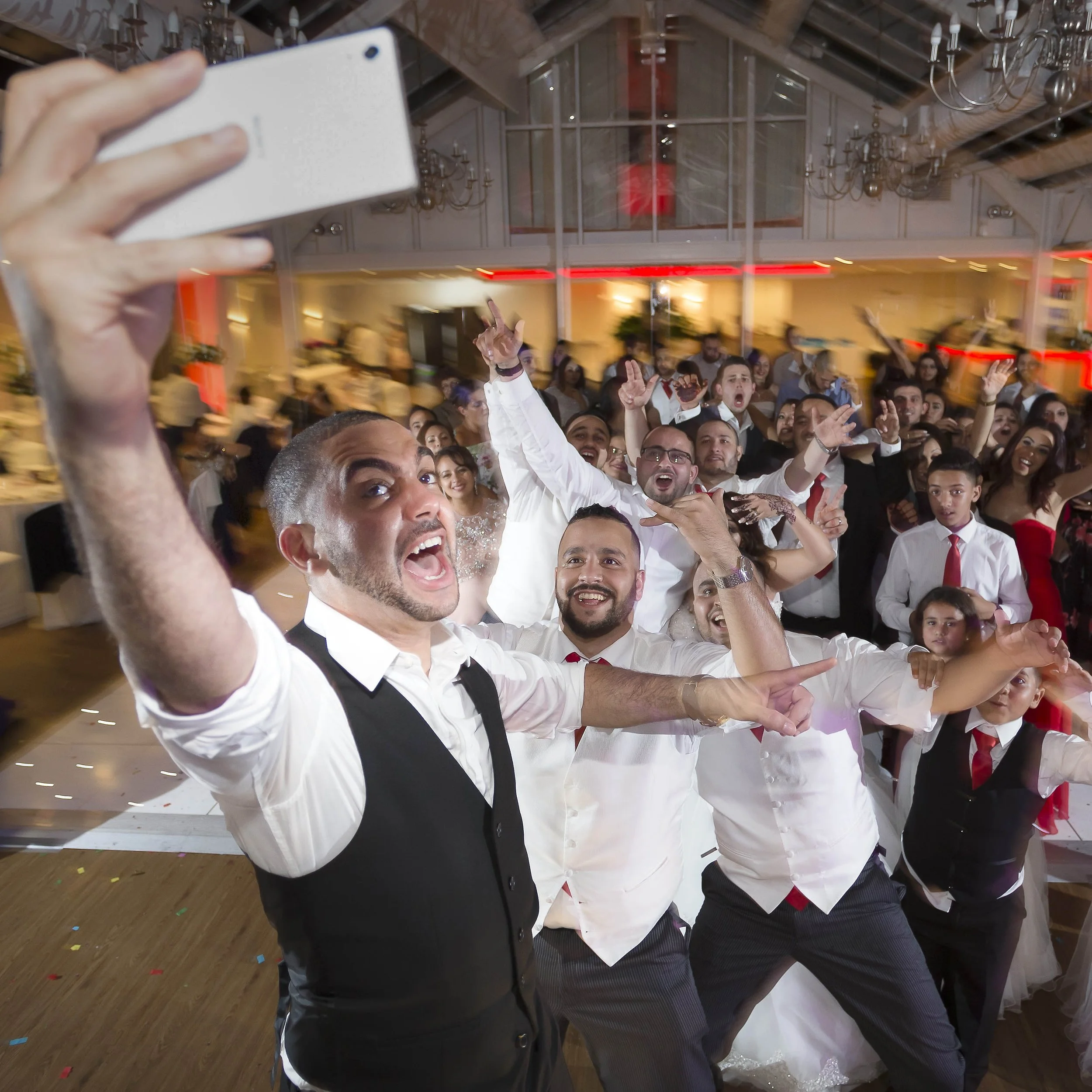 A group of people at a wedding reception taking a selfie, dancing, and celebrating indoors with chandeliers and warm lighting.