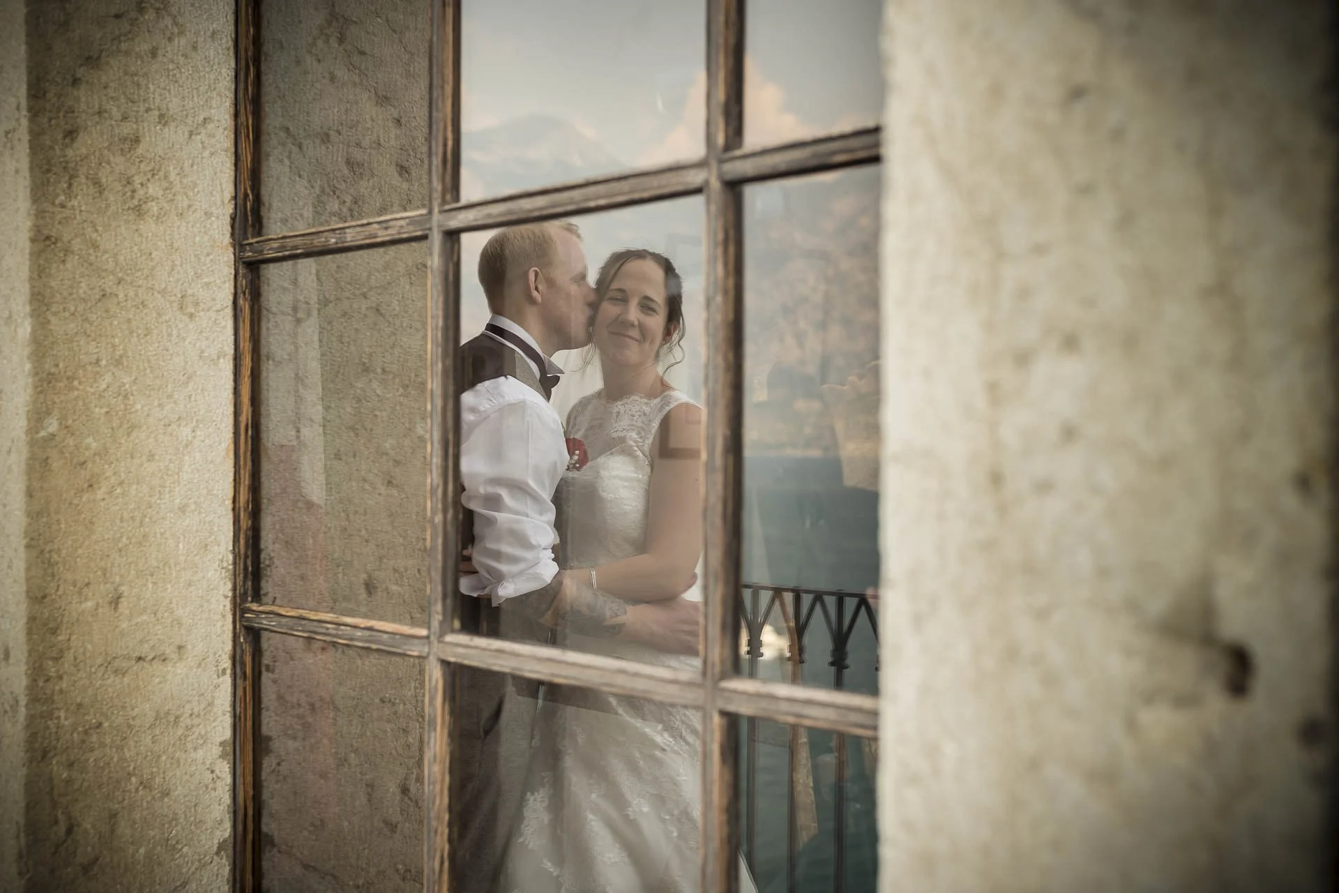 A bride and groom standing close together, kissing, seen through a window with rusted metal frame. The bride is smiling and wearing a white wedding dress, while the groom is dressed in a vest and shirt.