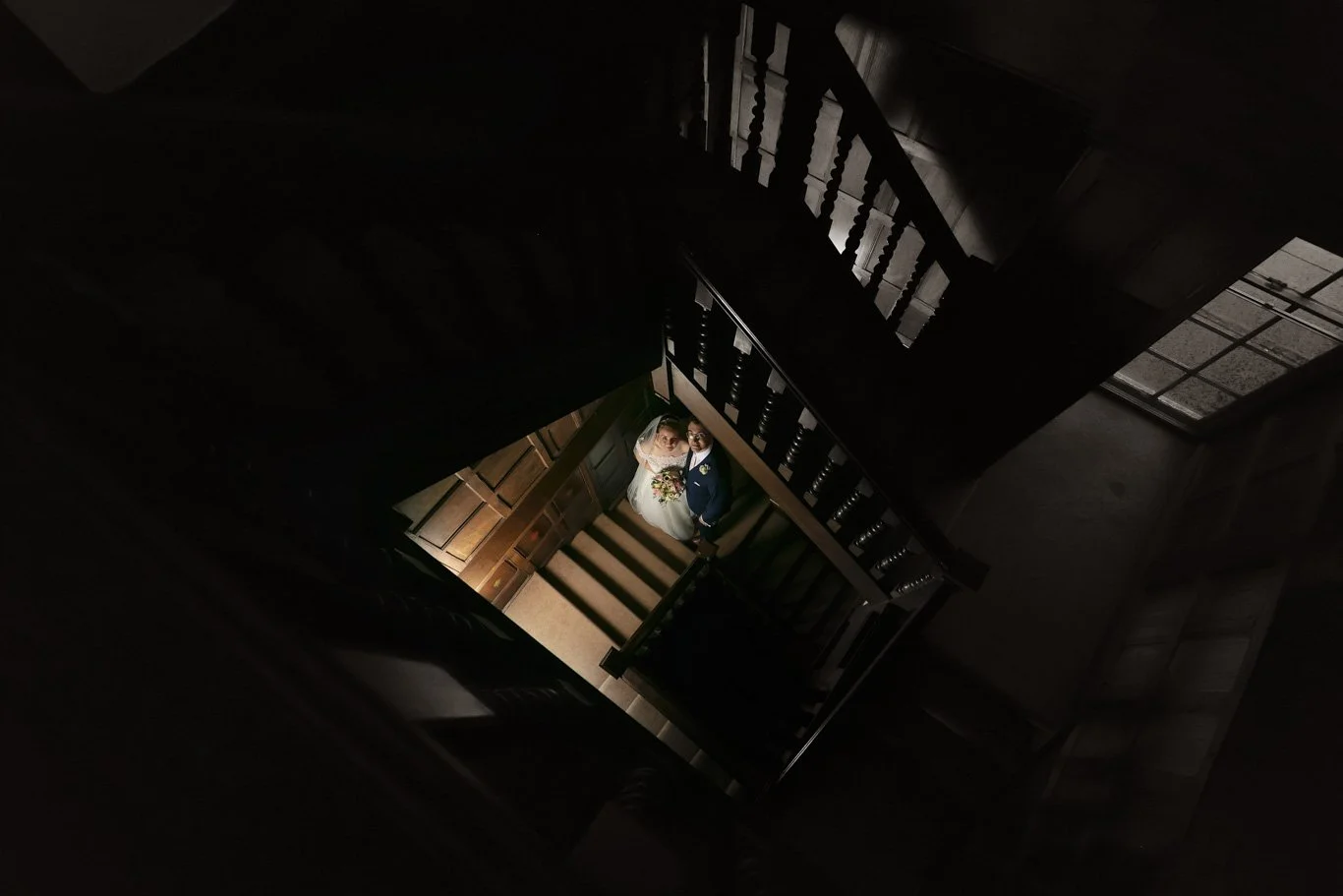 Looking down a staircase at a bride and groom standing together, illuminated in a dark interior, with wooden railings and stairs.