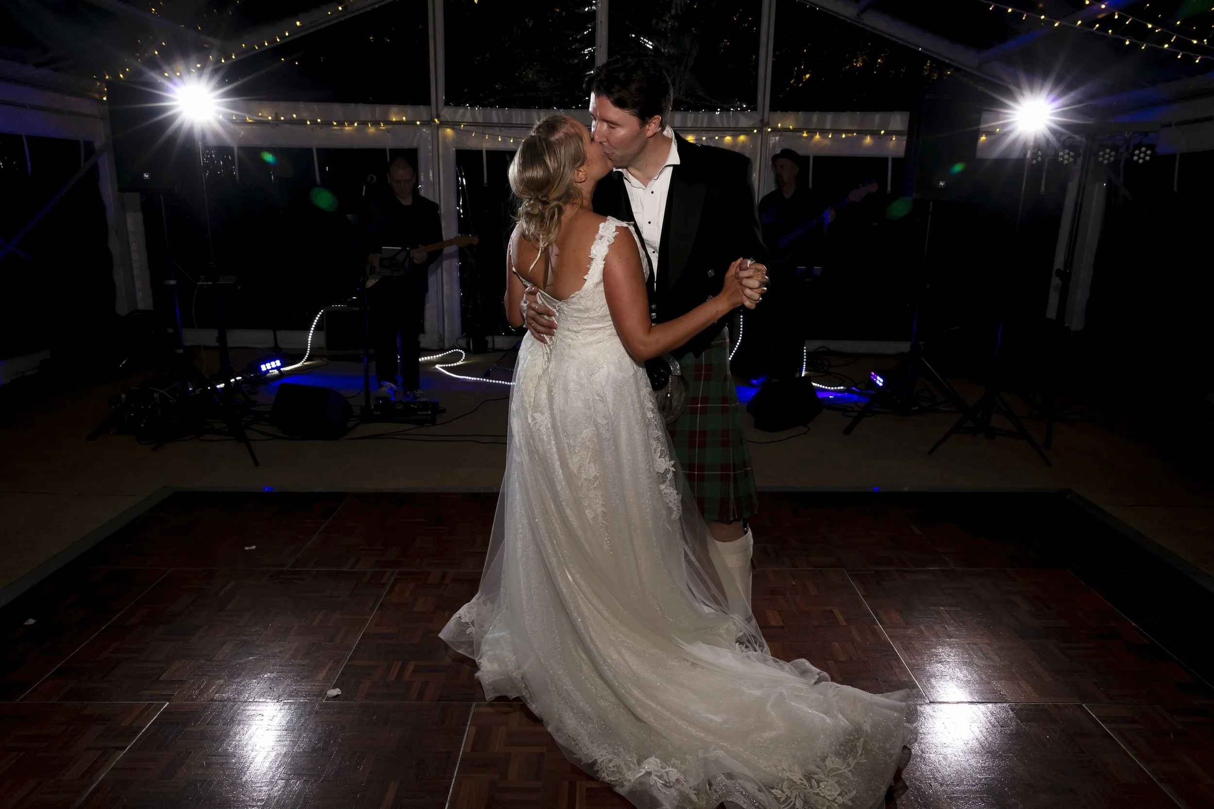A bride and groom dancing closely at their wedding reception in a decorated indoor venue with string lights and band members in the background.