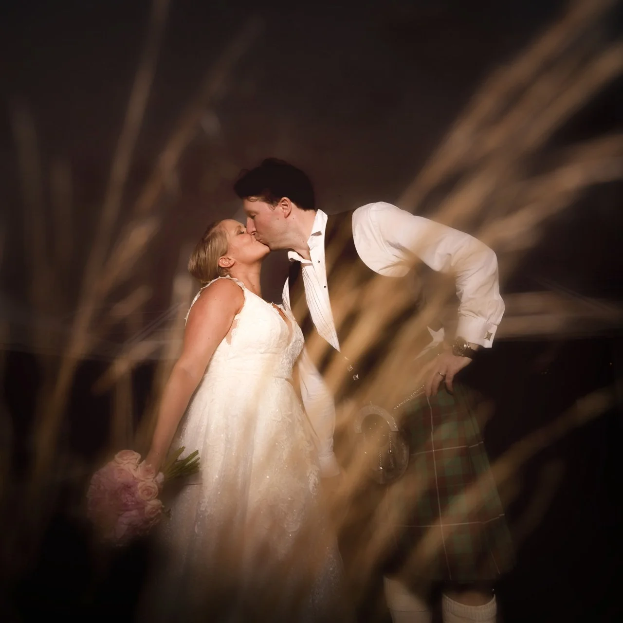 A bride and groom kissing on their wedding night, with blurred wheat stalks in the foreground and dark background.