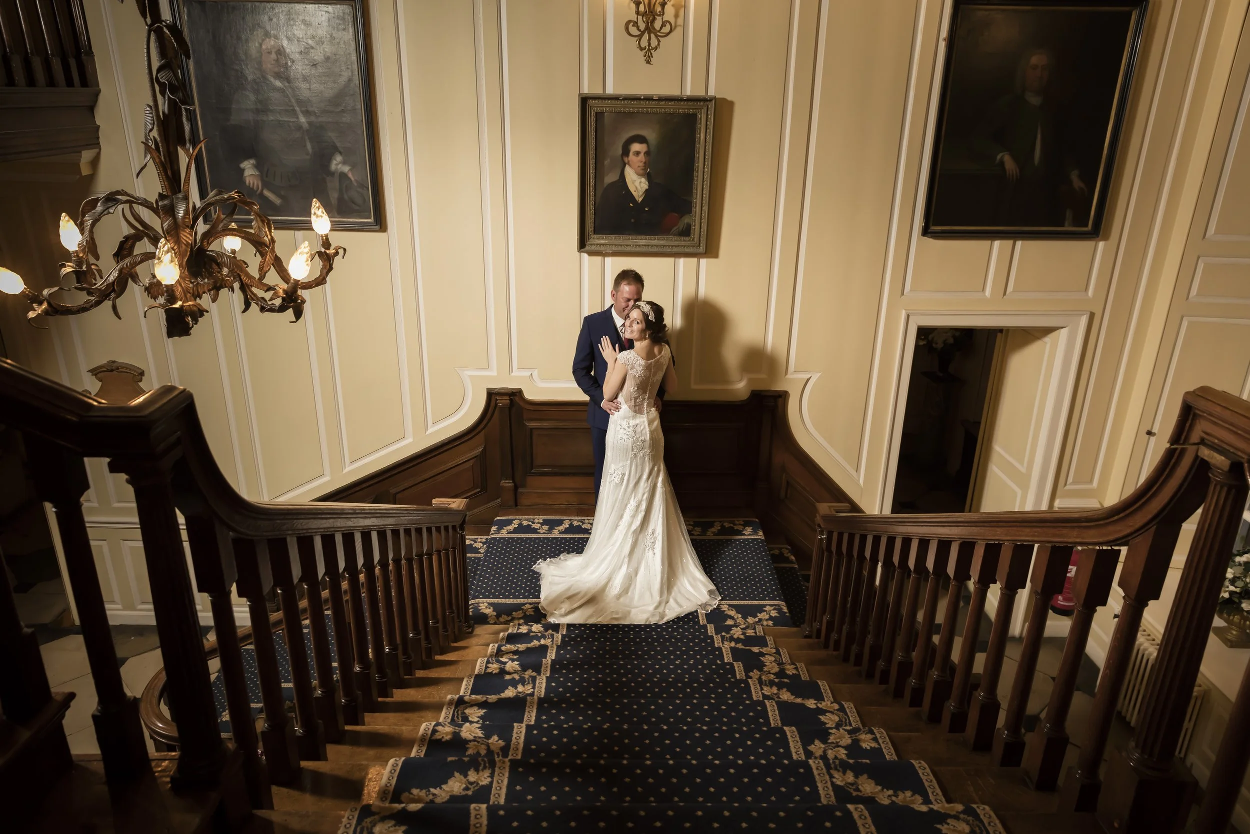A bride and groom embrace on a staircase in an elegant, historic building, with portraits on the wall behind them and ornate lighting.