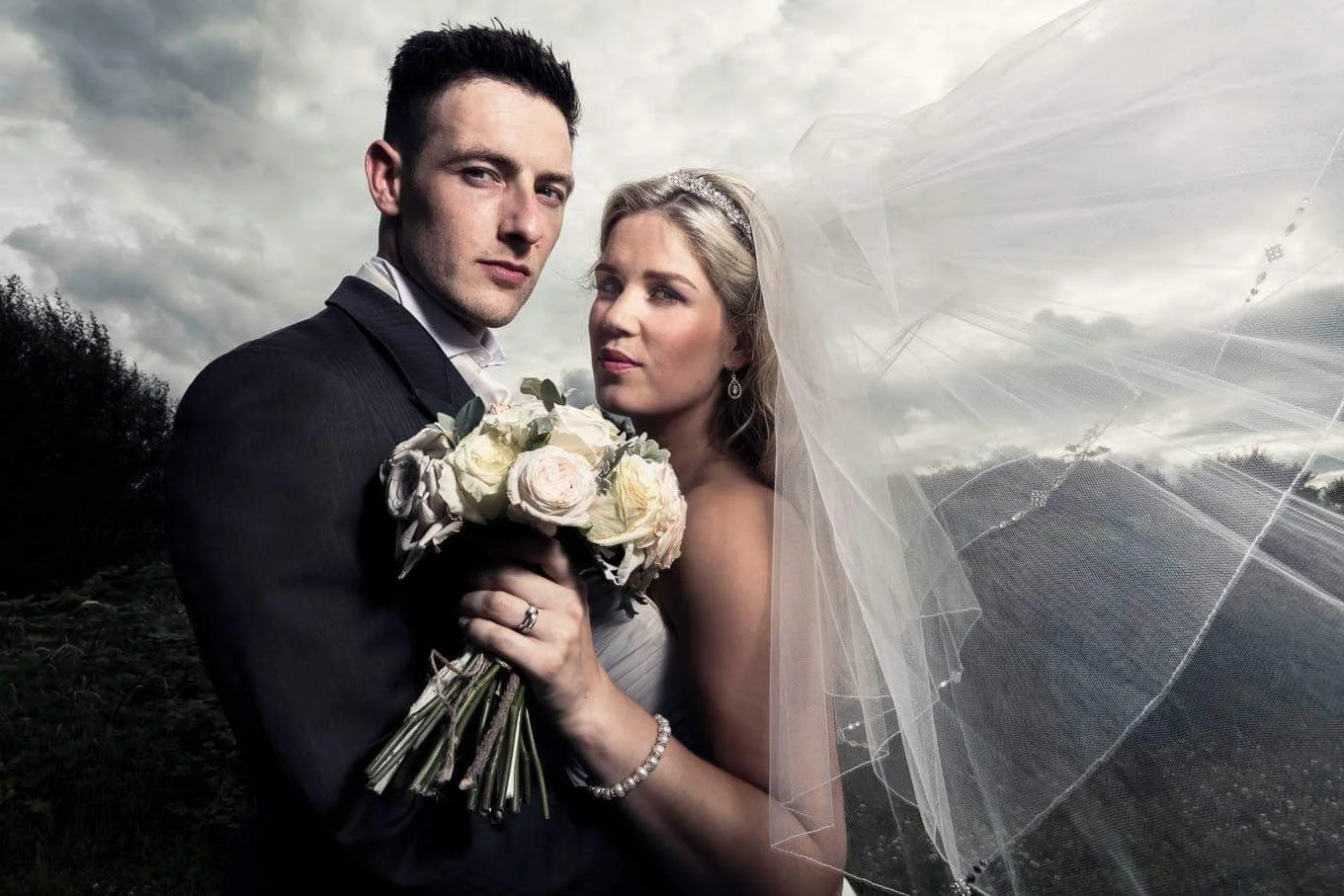 A newlywed couple standing outdoors against a cloudy sky, with the groom in a dark suit holding a bouquet of white roses and the bride in a wedding dress and veil, posing closely together.
