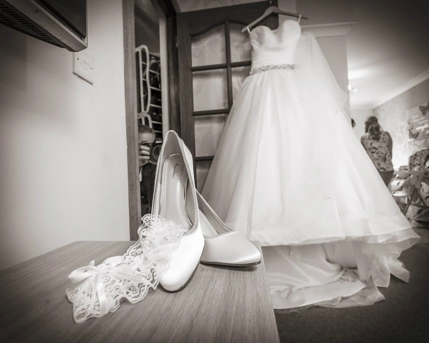 A pair of white bridal shoes with lace and satin ribbons on a wooden surface, with a wedding dress hanging in the background.