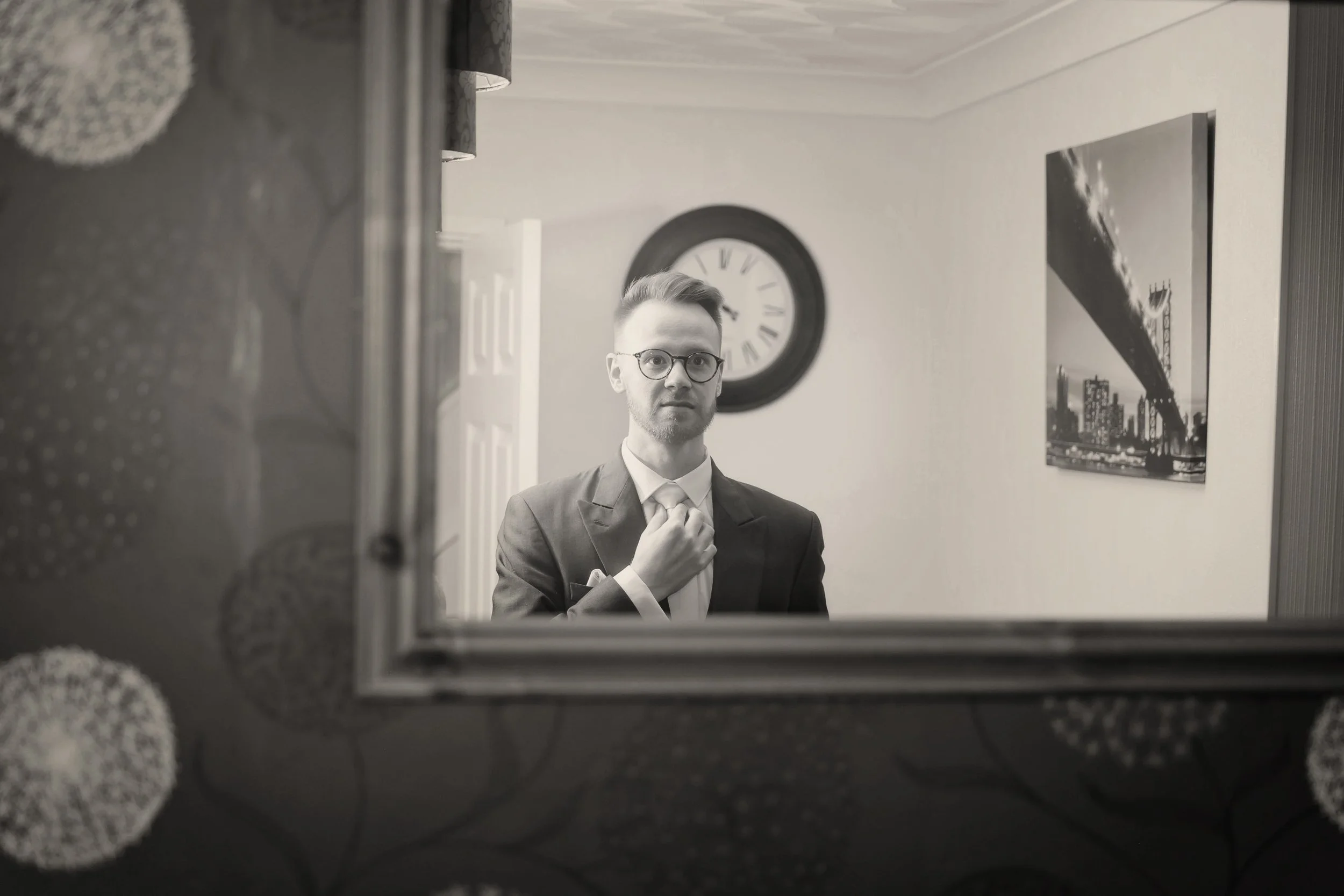 A young man in a suit and glasses looking into a mirror, adjusting his tie, with a clock and a cityscape photograph on the wall behind him.