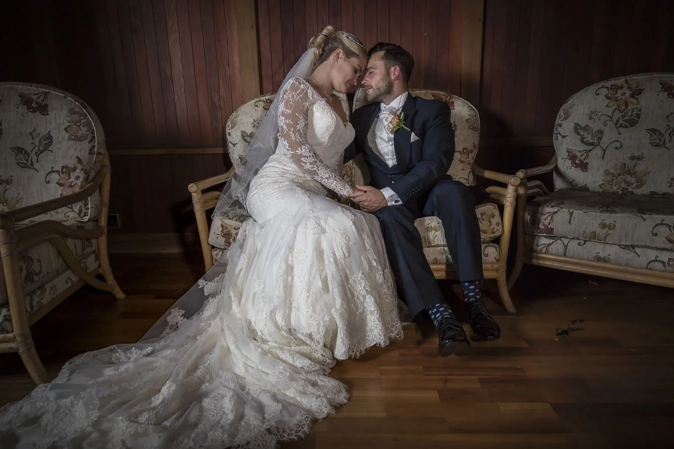 A bride and groom sitting closely on a floral-patterned couch, holding hands, with foreheads touching in a tender moment, in an intimate indoor setting.