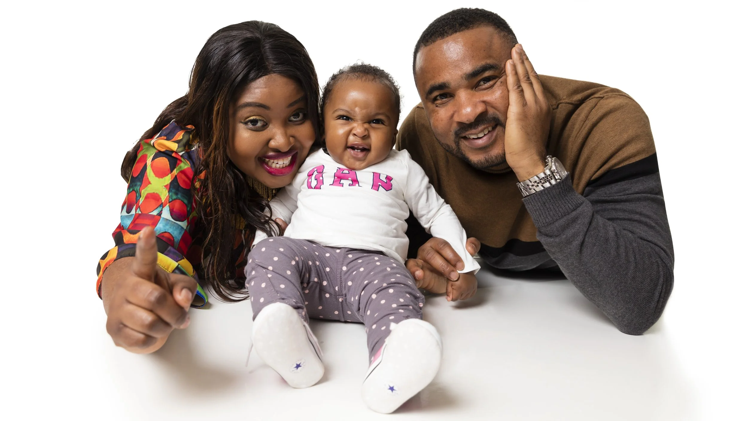 A happy family of three lying on the floor, smiling and making silly faces. The woman on the left has dark hair and is wearing a colorful patterned shirt, giving a thumbs-up. The baby girl in the middle is wearing a white shirt with pink letters and gray polka-dotted pants, and is making a playful face. The man on the right has short hair and is wearing a brown and gray sweater, resting his face on his hand.